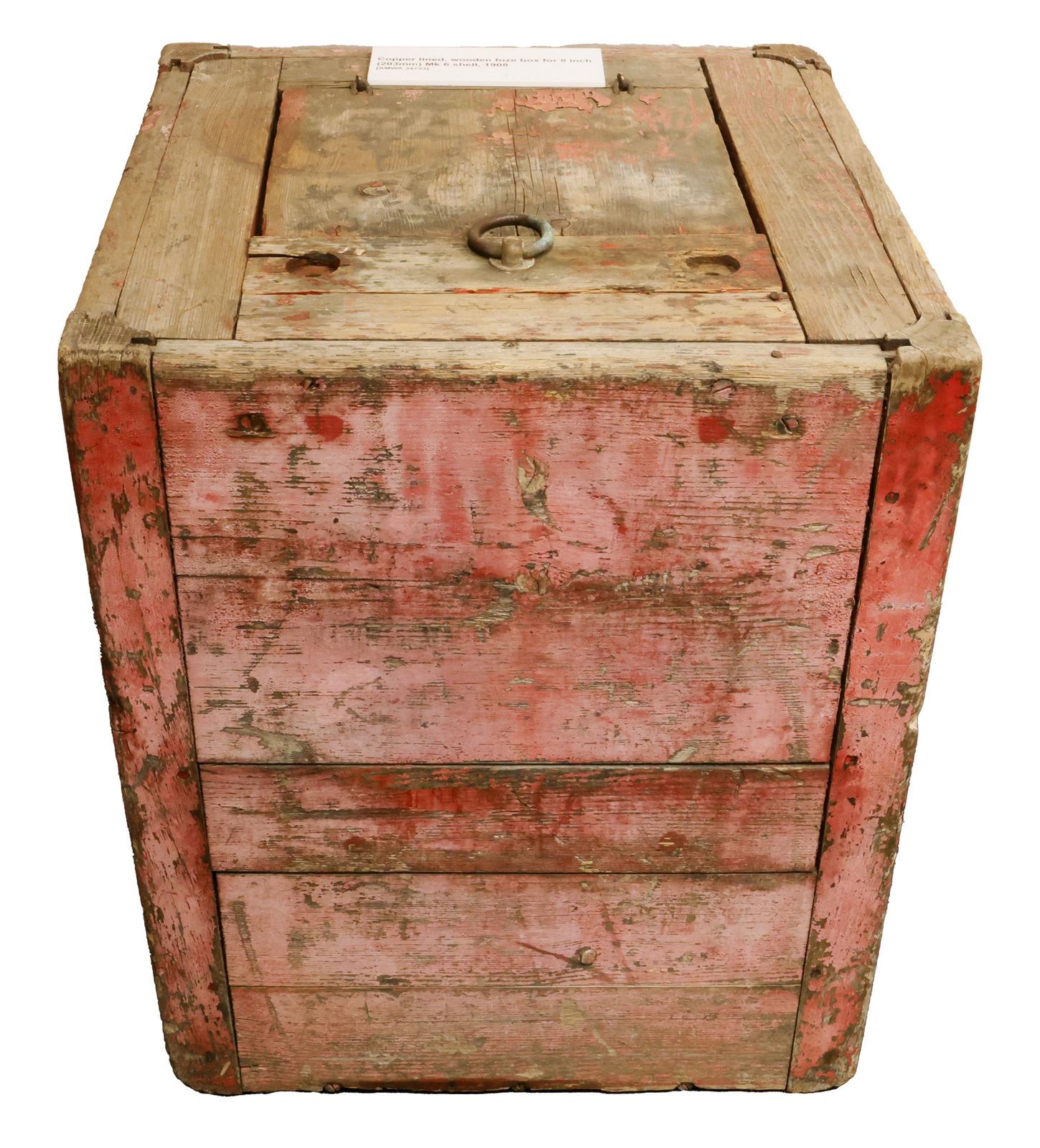 The image shows a weathered, wooden crate with a rustic, aged appearance. The crate is roughly cubic in shape, with visible wear and tear on the surface. The wood has a reddish tint, possibly from old paint or natural aging. There is a circular metal latch or knob on the top of the crate, indicating it may have been used for storage or shipping. The crate's surface appears rough and worn, with some visible cracks and imperfections.