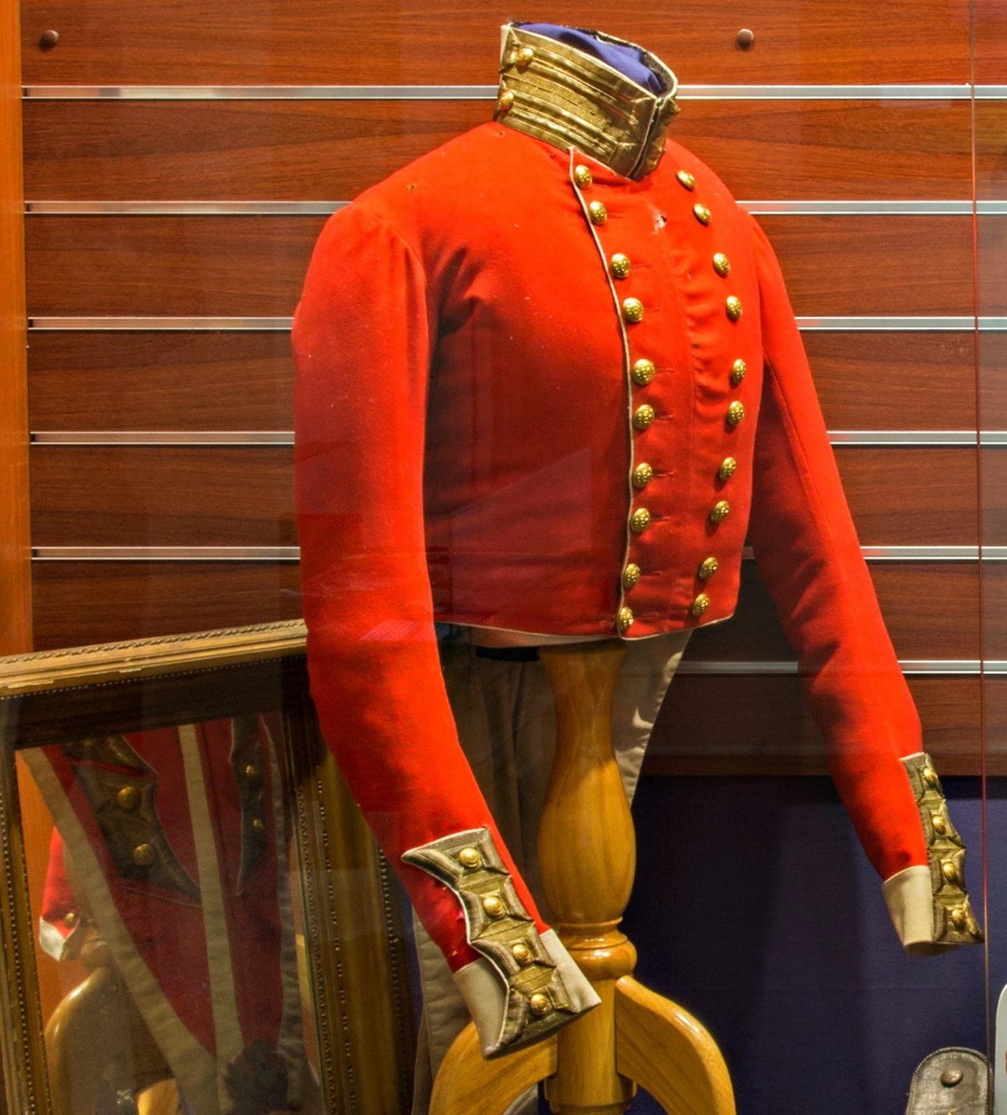 A historical red military jacket displayed on a wooden mannequin inside a glass case. The jacket has gold buttons arranged in two parallel rows down the front and ornate gold detailing on the cuffs and collar. The jacket is set against a wooden background with a reflection of another framed item visible in the lower left corner. This piece appears to be from an 1840s era military uniform.