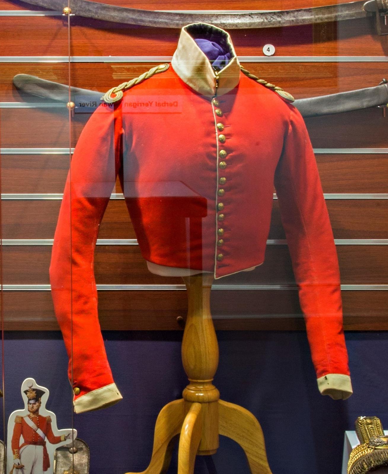 A red historical military jacket displayed on a wooden mannequin inside a glass case. The jacket features gold buttons running down the front, gold braided shoulder detailing, and a beige collar. A curved sword is mounted on the wall behind the jacket. To the bottom left, there is a small illustration of a soldier wearing a similar uniform. This jacket appears to be part of a 1840s military collection.