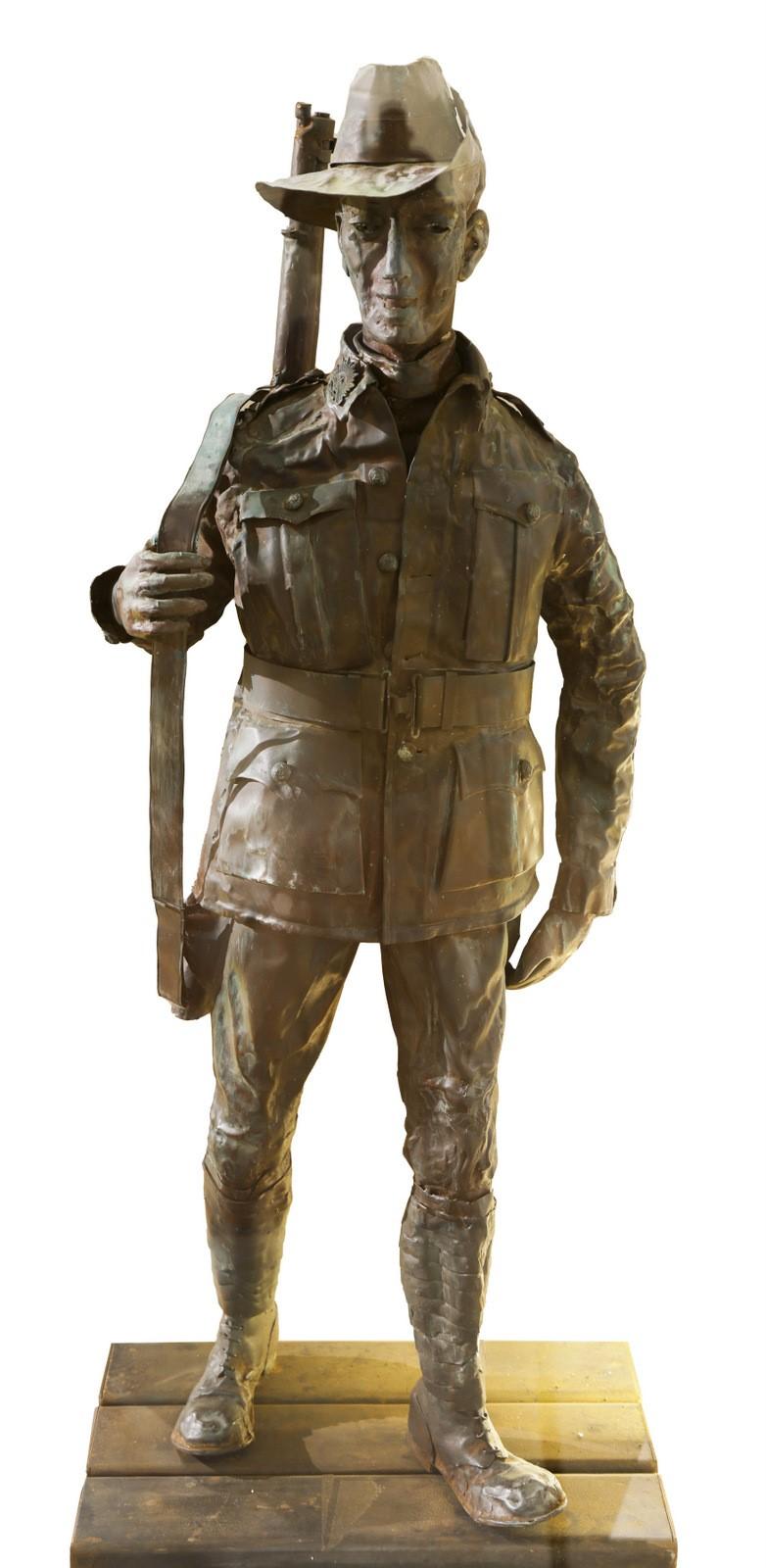 This image shows a copper statue of a soldier, often referred to as a "Digger," which is a term used to describe Australian soldiers, particularly from World War I and II. The soldier is wearing a slouch hat, a long military jacket with pockets, and boots. He holds a rifle resting on his right shoulder, with his hand gripping the barrel. The statue stands on a wooden platform, and the soldier's facial expression appears solemn and reflective. The detailing on the uniform, including buttons and the texture o