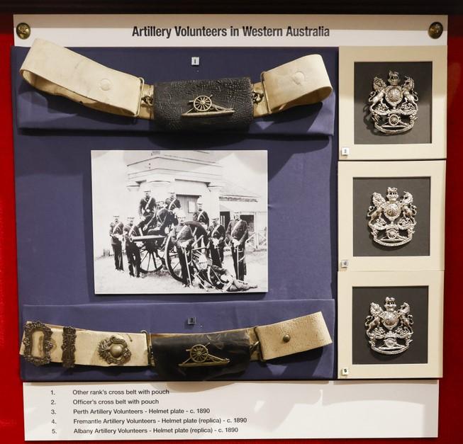 Display of historical artifacts titled 'Artillery Volunteers in Western Australia.' The exhibit showcases two white belts with metal buckles featuring artillery insignia, one at the top and one at the bottom of the display. In the center is a black-and-white photograph of artillery personnel and equipment. On the right side, there are three close-up images of artillery insignia. Below the display, a description lists the items, including belt buckles, buttons, and badges associated with Western Australia.