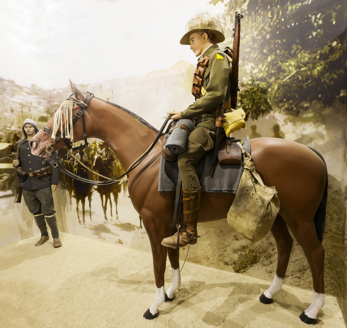 This image shows a life-size museum display of a soldier mounted on a horse, likely representing a scene from World War I or an earlier military period. The soldier is dressed in historical military attire, wearing a khaki uniform with a helmet, a rifle strapped across his back, and holding the reins of the horse. The horse is equipped with saddlebags and military gear. In the background, another figure in uniform stands beside the scene, suggesting a military or wartime setting. The backdrop appears to dep