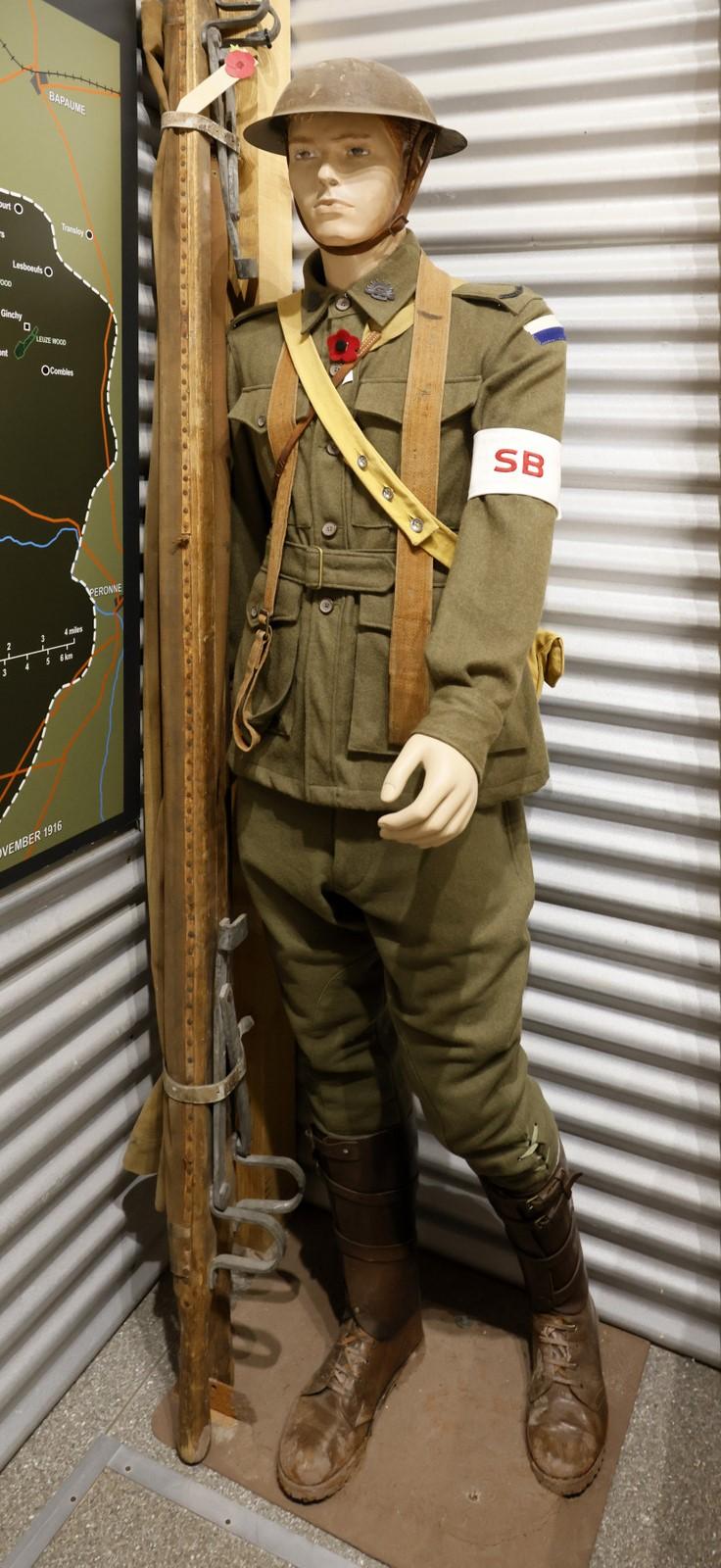  Image shows a life-size mannequin dressed in a World War I British soldier uniform. The soldier wears a khaki-colored uniform, including a tunic, trousers, and boots, along with a metal helmet. The mannequin is equipped with a rifle propped upright next to it, with a bayonet attached. The soldier also wears a white armband with a red cross, indicating a medic role. There is a brown leather belt, pouches, and a backpack, with various items of military gear attached. The display appears to be part of a mus