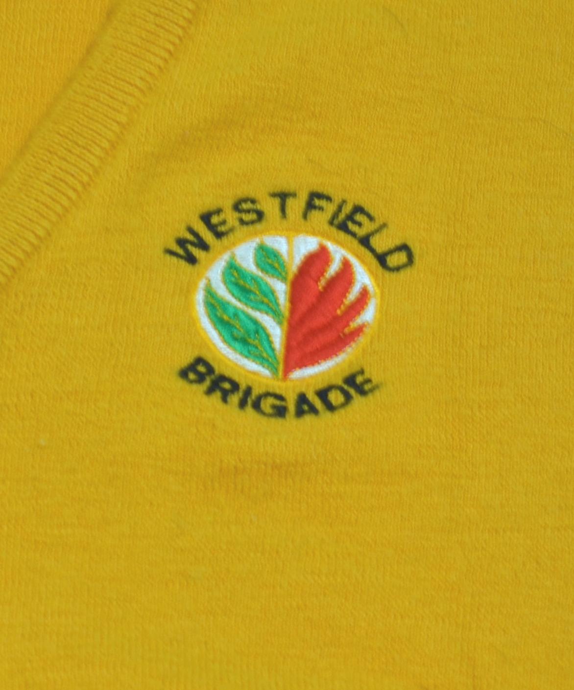 A logo on a yellow woolen jumper. Embroidered on the left chest is a white circle with stylised green leaves on the left and red flames on the right. In a circle around the logo are the words WESTFIELD and BRIGADE in black cotton.