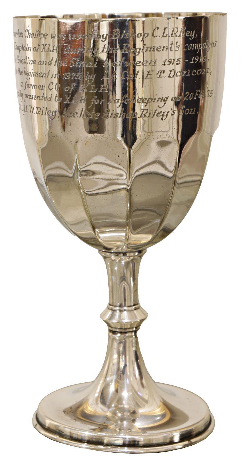 A silver chalice with a polished surface, engraved with text around the upper part of the cup. It has a classic goblet shape with a round base, slender stem, and a wide bowl at the top. The chalice reflects light, highlighting its metallic sheen.