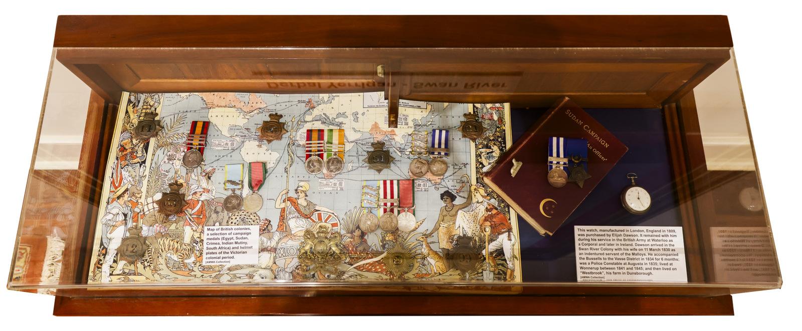 A display case containing various military medals and decorations arranged on a map background. The medals are placed at different points on the map, possibly indicating locations of significance. To the right, there is a blue plaque or document and an additional medal, along with an information card. The display case has a wooden frame with a glass cover.