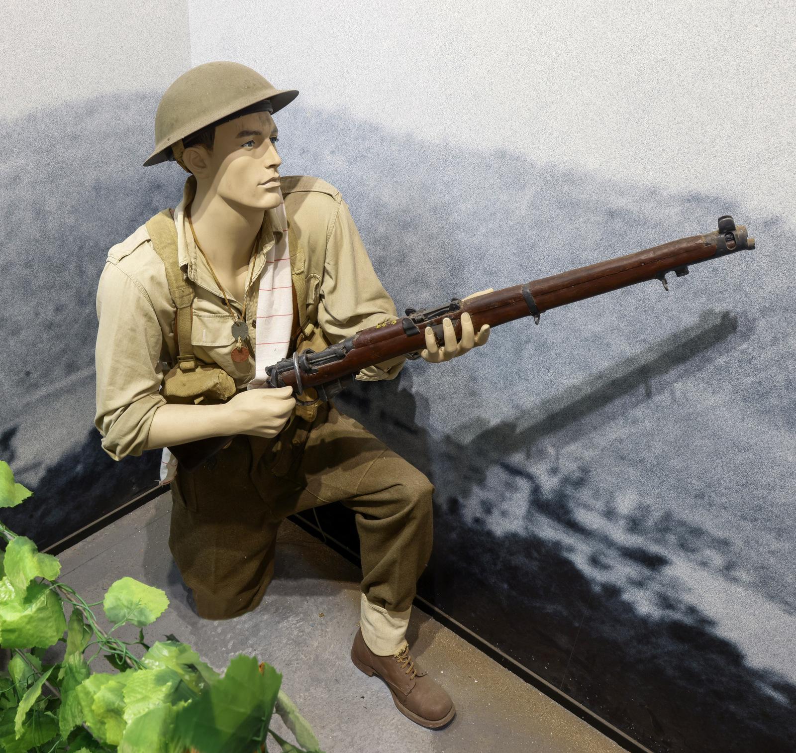 Mannequin of a World War II soldier wearing a helmet and khaki uniform, kneeling and aiming a vintage rifle. The display is set against a backdrop of a black-and-white wartime scene, with greenery in the foreground