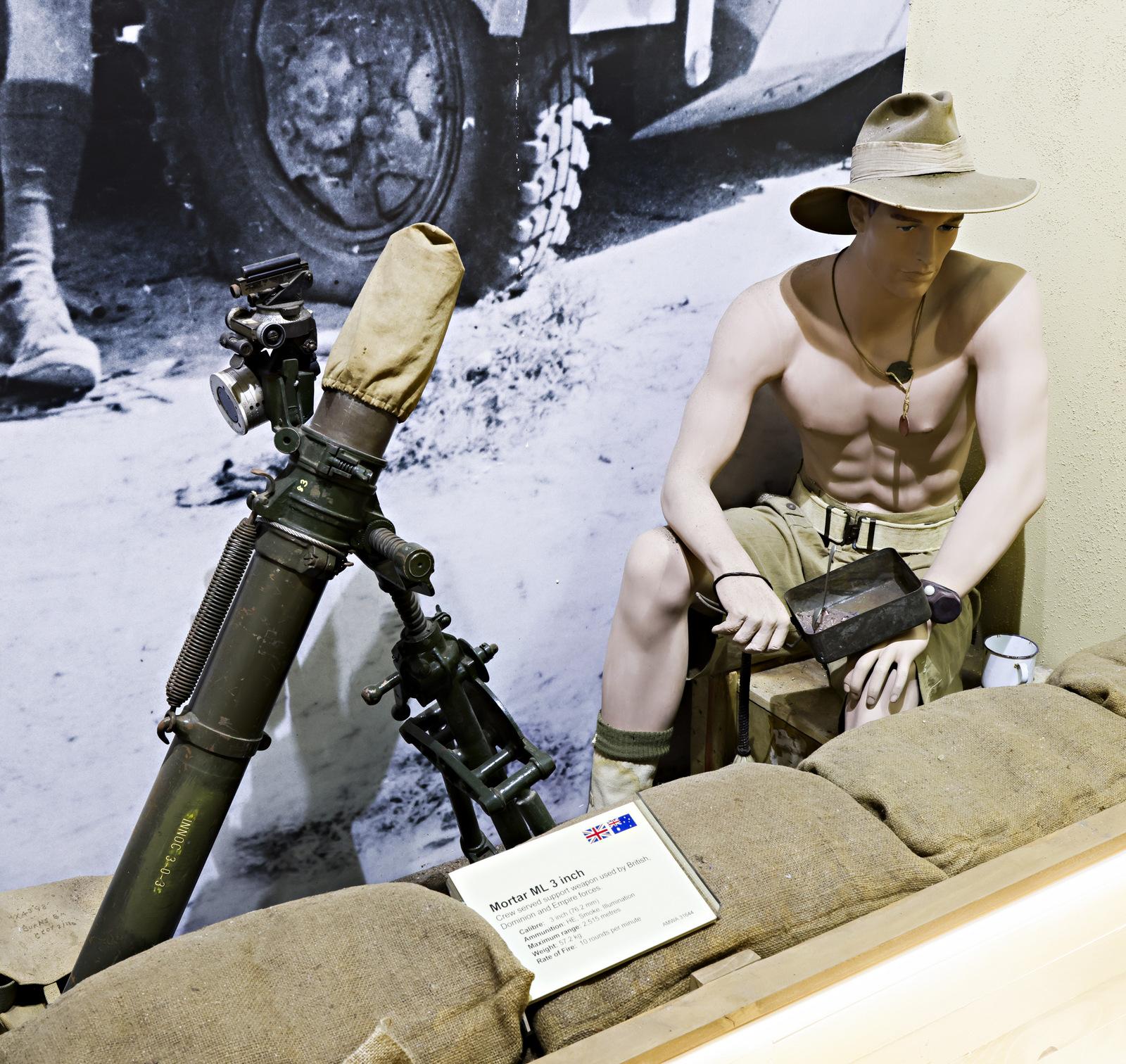 A museum exhibit showcasing a vintage military mortar setup, with a mannequin dressed as a shirtless soldier wearing a wide-brimmed hat and shorts, sitting beside the mortar. The display is accompanied by sandbags, a descriptive placard, and a black-and-white wartime photograph in the background.