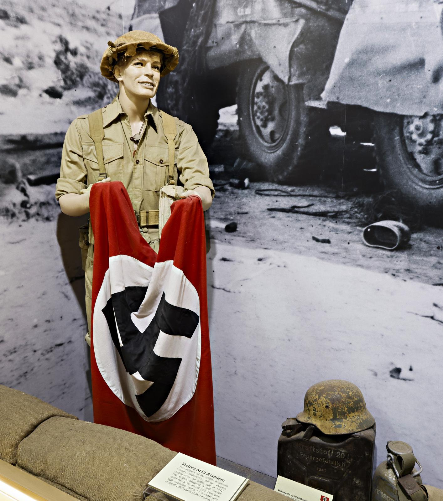 Mannequin dressed as a World War II soldier holding a Nazi flag, displayed in a museum exhibit with historical artifacts, including a helmet and jerrycan, against a backdrop of wartime imagery.