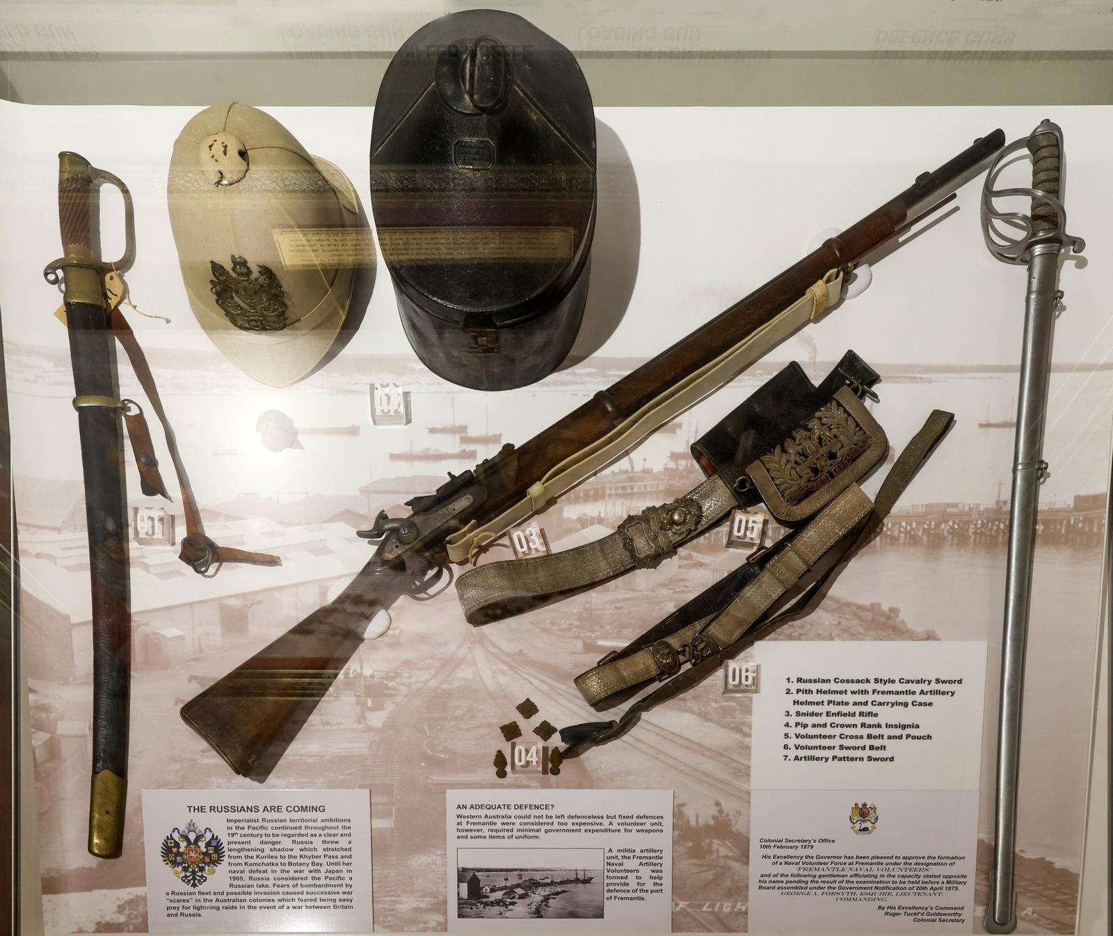 A museum display showcasing historical military equipment, including two hats, a rifle, two swords, a belt with ammunition pouches, and various small items. The display is set on a white background with informational placards providing descriptions of the artifacts. The items appear to be from a late19th-century military collection.