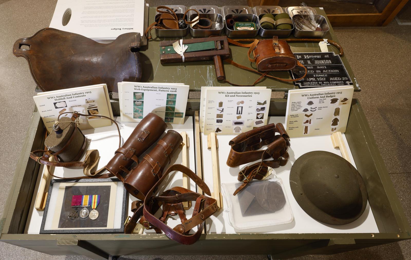 "A display of World War I-era military artifacts arranged on a table. The items include a leather satchel, binoculars in leather cases, various belts and pouches, a steel helmet, a collection of medals, insignia, and buttons, as well as documents explaining each item's significance. The layout provides a detailed look at the personal gear and insignia used by soldiers during the war."