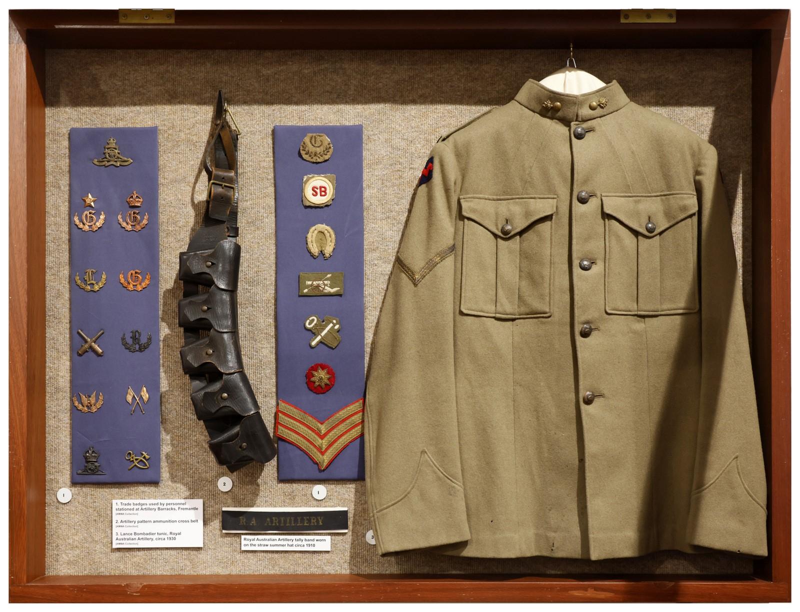 A display case containing a vintage military uniform and various insignia. The uniform is an olive-green jacket with two chest pockets, buttoned up and displayed to the right. To the left of the jacket are rows of military insignia, including pins, badges, and patches mounted on blue felt. An ammunition belt is displayed alongside the insignia. Each item is labeled with small descriptive plaques. The display case is framed in wood with a glass front.