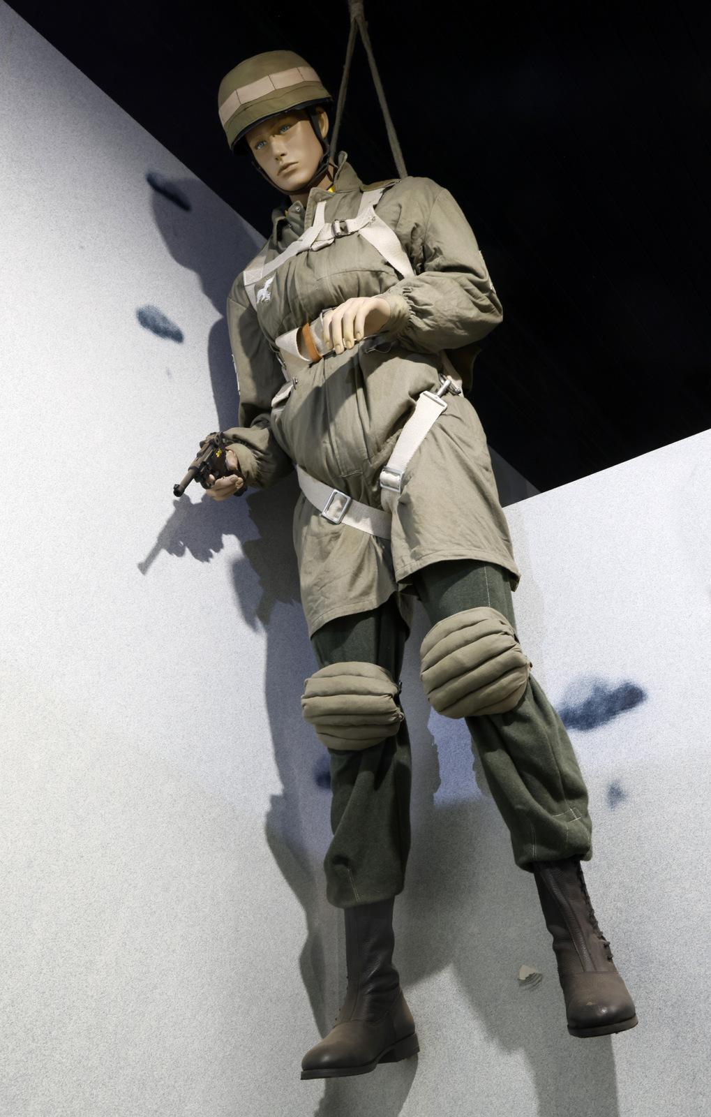 A mannequin dressed in vintage German military paratrooper gear is suspended in mid-air, simulating a descent. The figure is outfitted in a green uniform with knee pads, a helmet, and holding a small firearm in one hand. The mannequin is harnessed with straps attached to the ceiling, creating the appearance of a parachute jump.