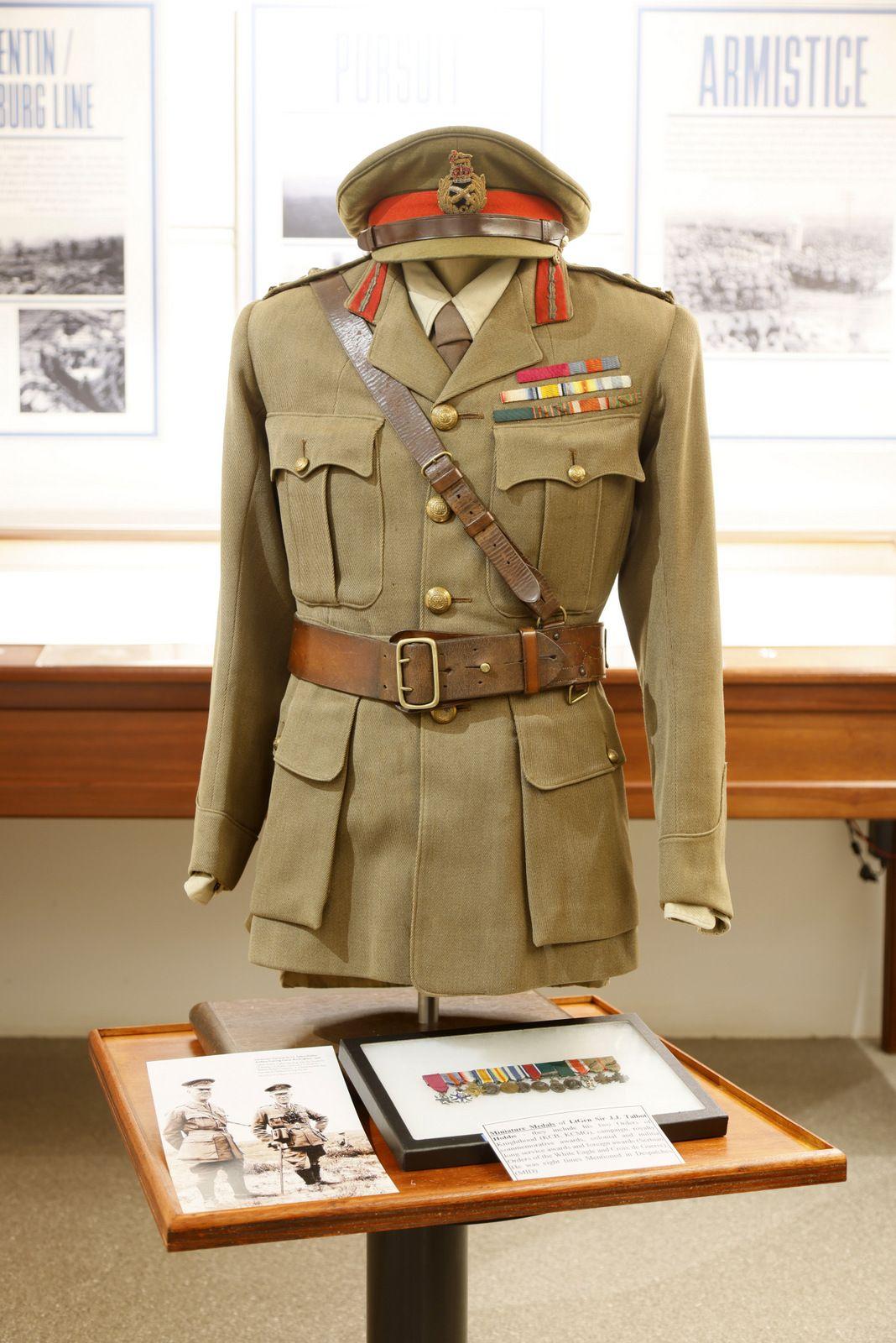 A military uniform displayed on a mannequin in a museum setting. The uniform consists of an olive-green jacket with red and gold insignia on the collar, a brown leather belt, and a matching hat. Medals and badges are visible on the chest. The display is on a wooden stand with informational booklets and photos placed on a table in front of the uniform. Historical posters are visible in the background