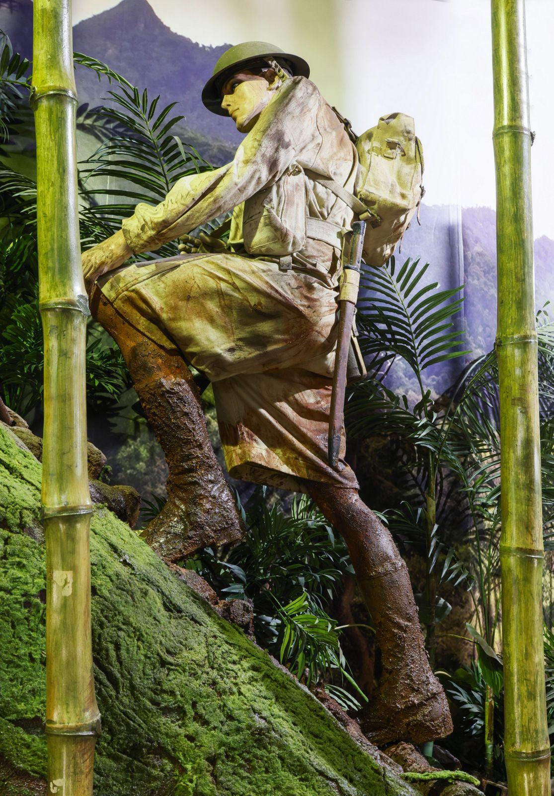 A life-sized sculpture of an Australian soldier dressed in khaki uniform and webbing, including a helmet khaki shirt, and boots, climbing a mossy hill. He carries a backpack and appears to be trekking through a lush jungle environment with bamboo stalks and dense greenery surrounding him. The scene suggests the conditions experienced on the Kokoda Trail in New Guinea