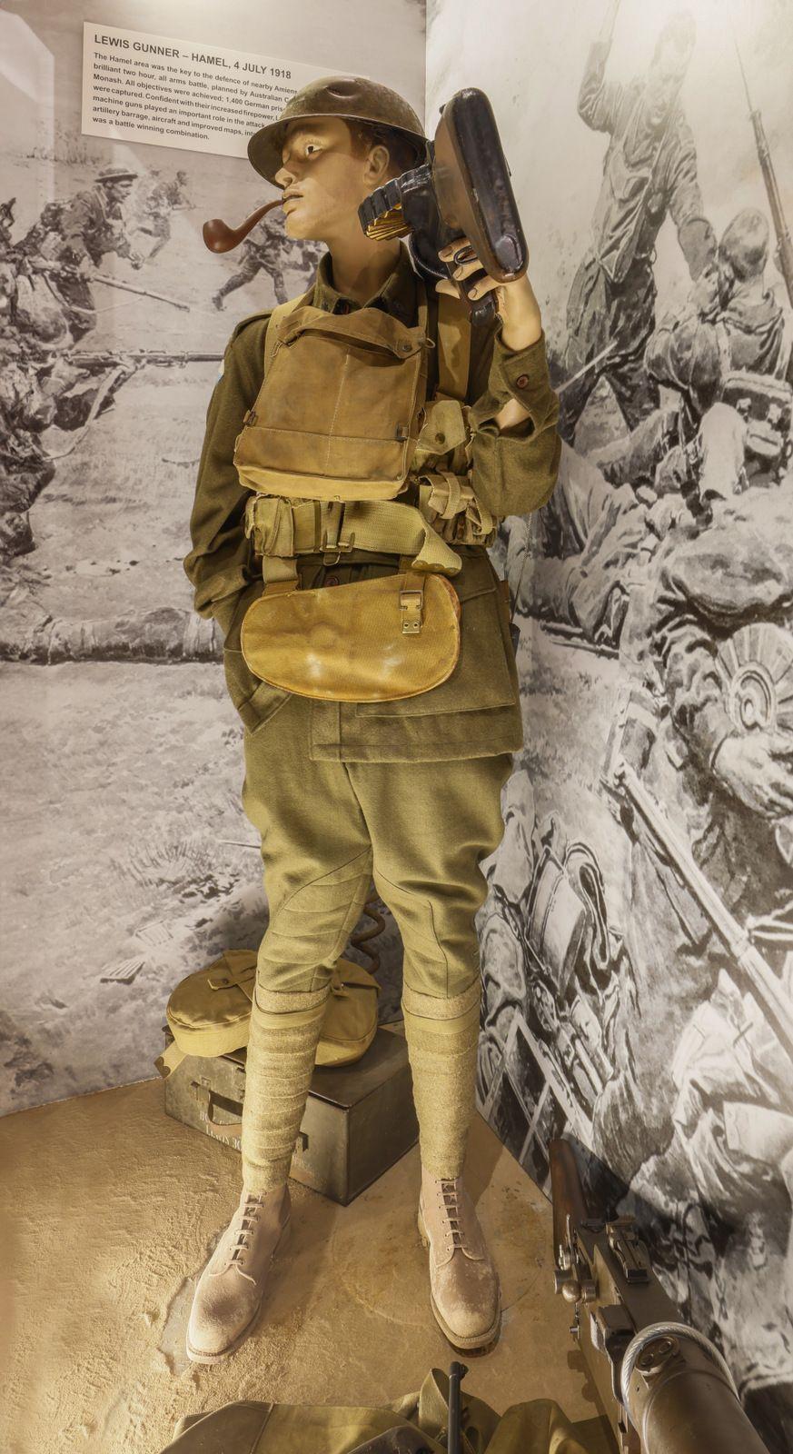 "A museum display of a World War I soldier mannequin dressed in period military uniform. The figure is wearing a helmet, carrying a Lewis gun on its back, and has various gear strapped to its body, including a vest and pouches. The backdrop shows a black-and-white image depicting a battlefield scene."