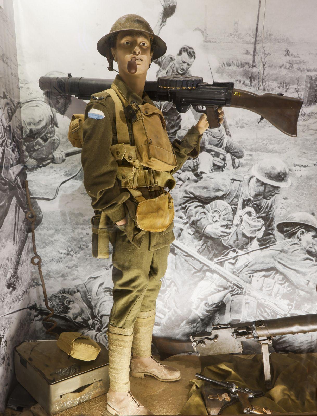 "A museum display of a World War I soldier mannequin dressed in period military uniform. The figure is wearing a helmet, carrying a Lewis gun on its back, and has various gear strapped to its body, including a vest and pouches. The backdrop shows a black-and-white image depicting a battlefield scene."