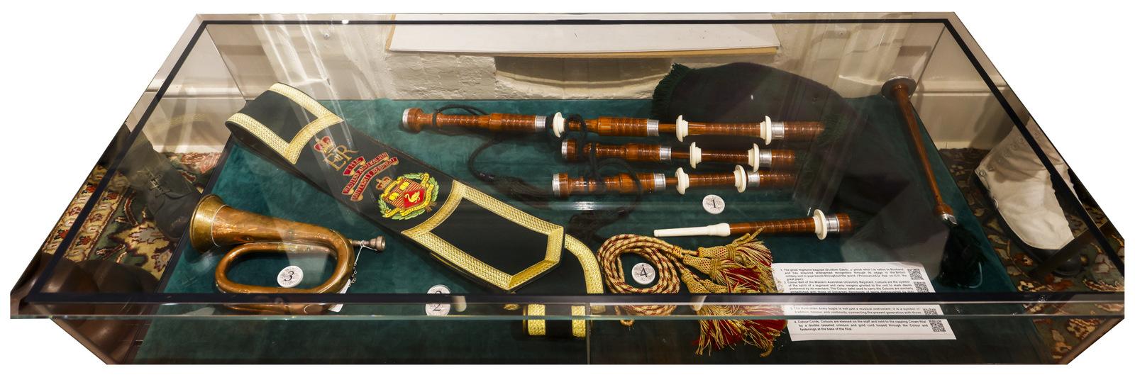  A display case containing a collection of historical musical instruments and accessories, including a brass instrument, bagpipes, and other decorative components. The items are arranged on a green felt base, with small identification tags visible.