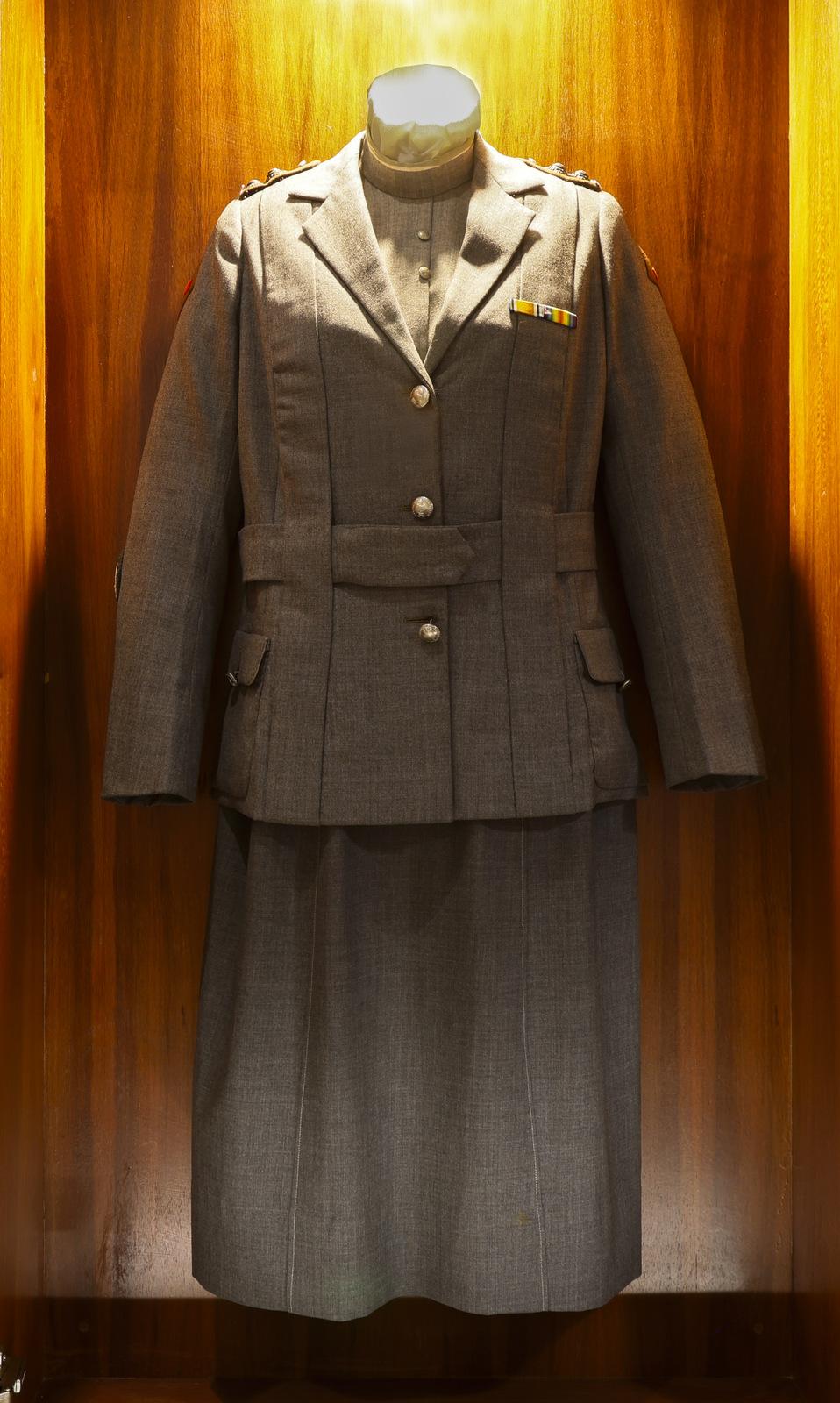 Photograph of a World War 1 Australian nurse's uniform displayed on a mannequin. The uniform consists of a grayish-brown tailored jacket with two front buttons and flap pockets, paired with a matching knee-length skirt. The background features a wooden panel, providing a warm and elegant contrast to the outfit.