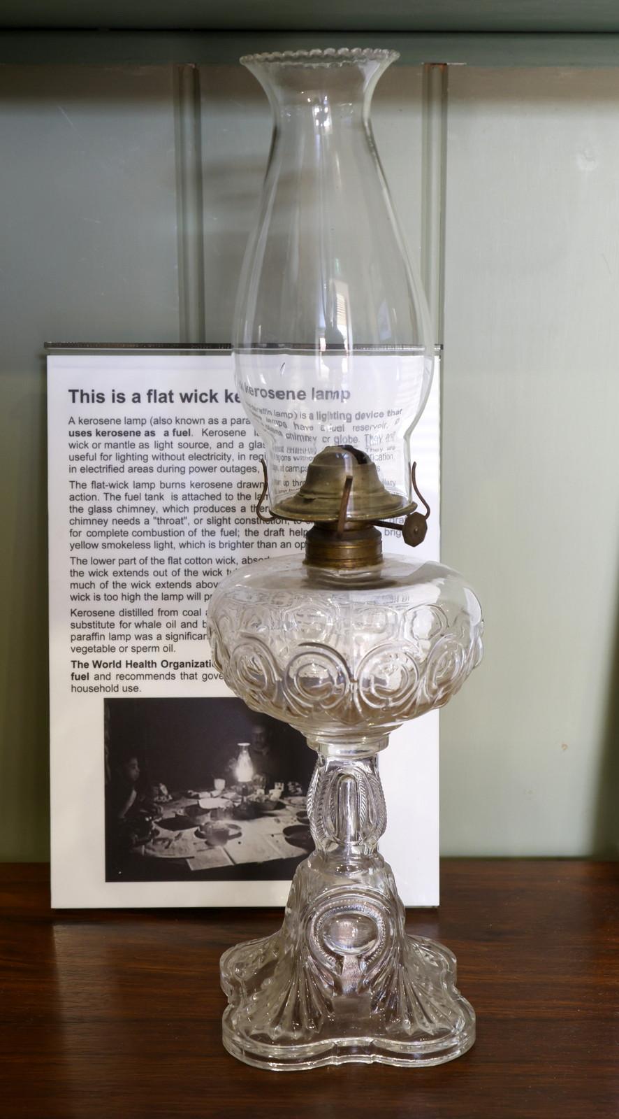 A vintage kerosene lamp with a decorative glass base and a tall, clear glass chimney. The lamp is displayed on a wooden surface with an informational sign placed behind it. The sign includes an image and text describing the history or details of the lamp.