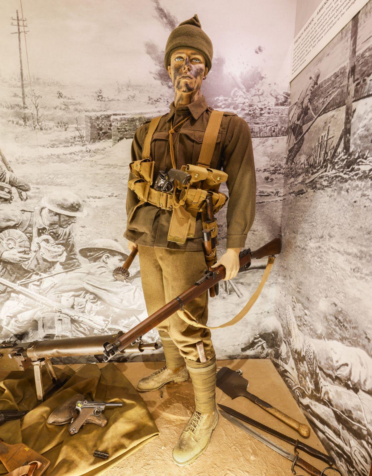 A museum exhibit displays a mannequin dressed as a World War I trench raider soldier. The soldier is equipped with a vintage rifle and wears a brown uniform with a woolen cap. The uniform includes a belt with various pouches for ammunition and equipment. The mannequin's face is painted to resemble dirt or camouflage, evoking the rugged conditions of trench warfare. At the mannequin's feet, various items are laid out, including a handgun, a shovel, a knife, and additional military gear. The background featur