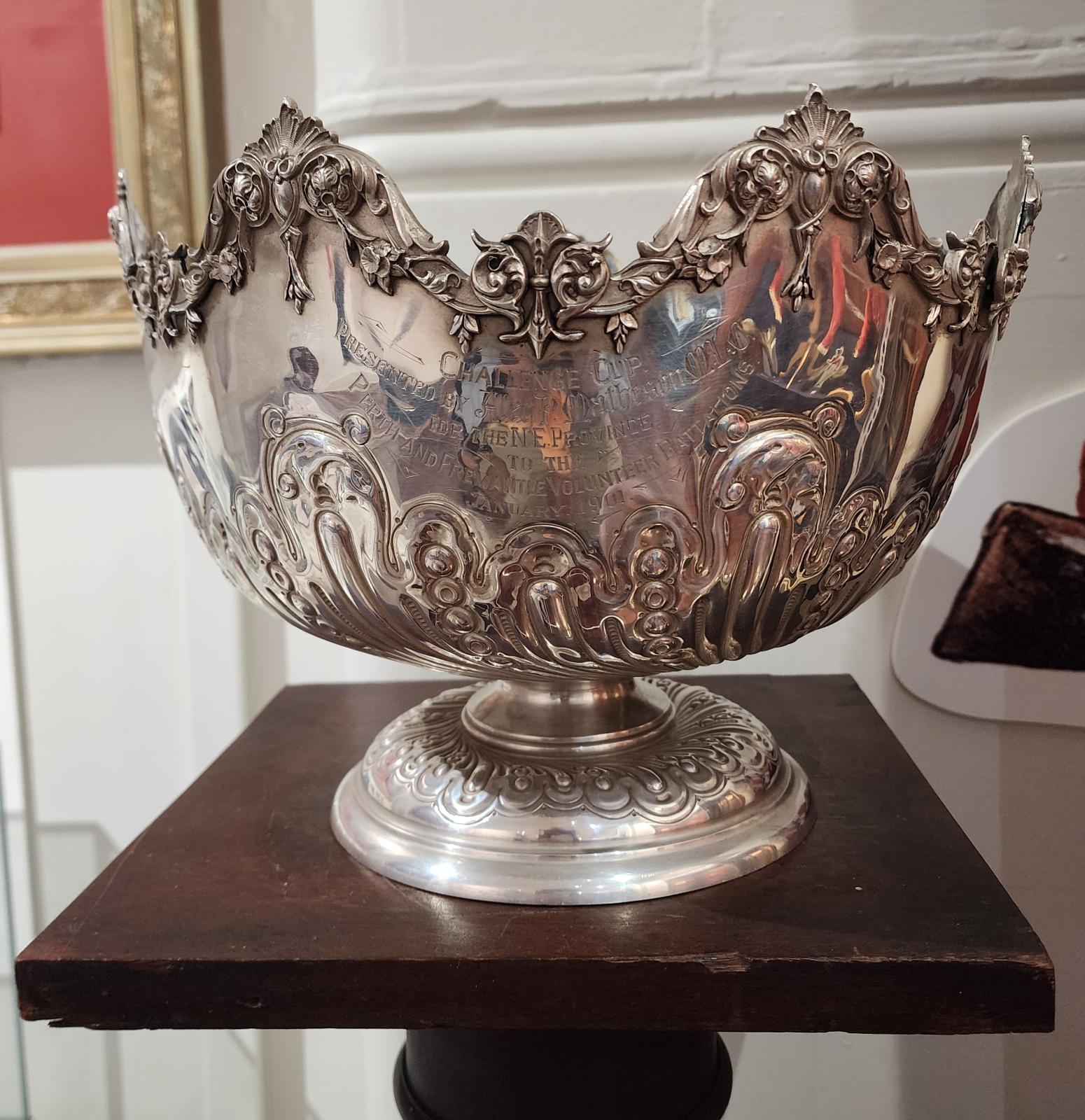 An ornate silver bowl on a pedestal, featuring an intricate design. The bowl has a scalloped rim adorned with floral and decorative elements. The body of the bowl is embossed with intricate patterns, including floral and possibly heraldic designs. The bowl sits on a wide, round base with additional detailed engravings. The piece has a reflective finish, showing its polished metal surface.