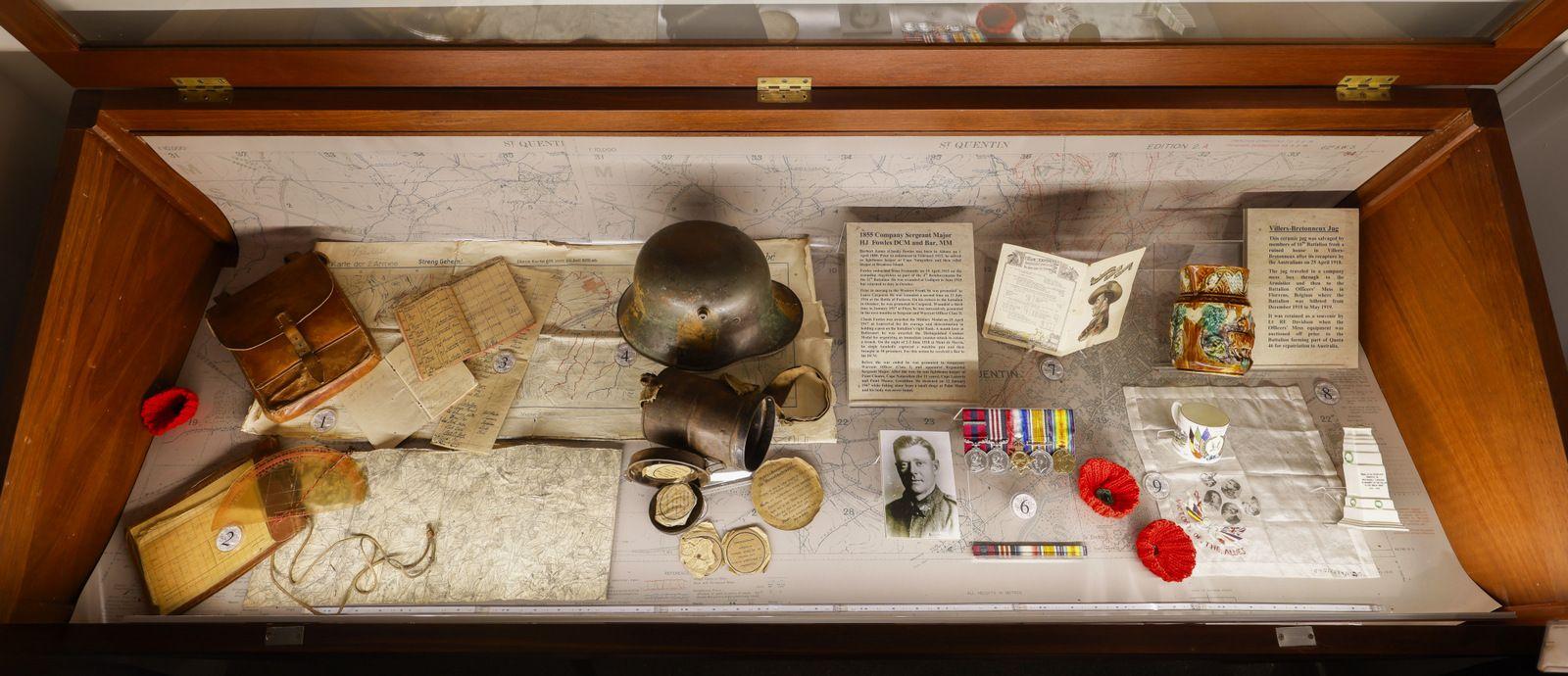 The image shows a display case containing various historical artifacts and documents. The items include old books, handwritten letters, maps, a metal helmet, a gas mask, small containers, medals, red poppies, and other memorabilia. The arrangement suggests a collection related to wartime or military history, presented on a light background with a protective glass cover.