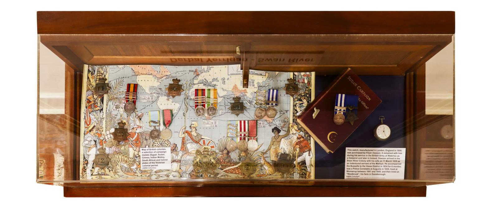 This is an image of a display case containing various military medals and decorations. The items are arranged on a map backdrop, and a small book or document is included on the right side. The case also contains a descriptive plaque, although the text is not legible in the image. The medals appear to be historical or commemorative in nature, signifying achievements or honors. The wood frame of the case gives it an elegant, preserved appearance.