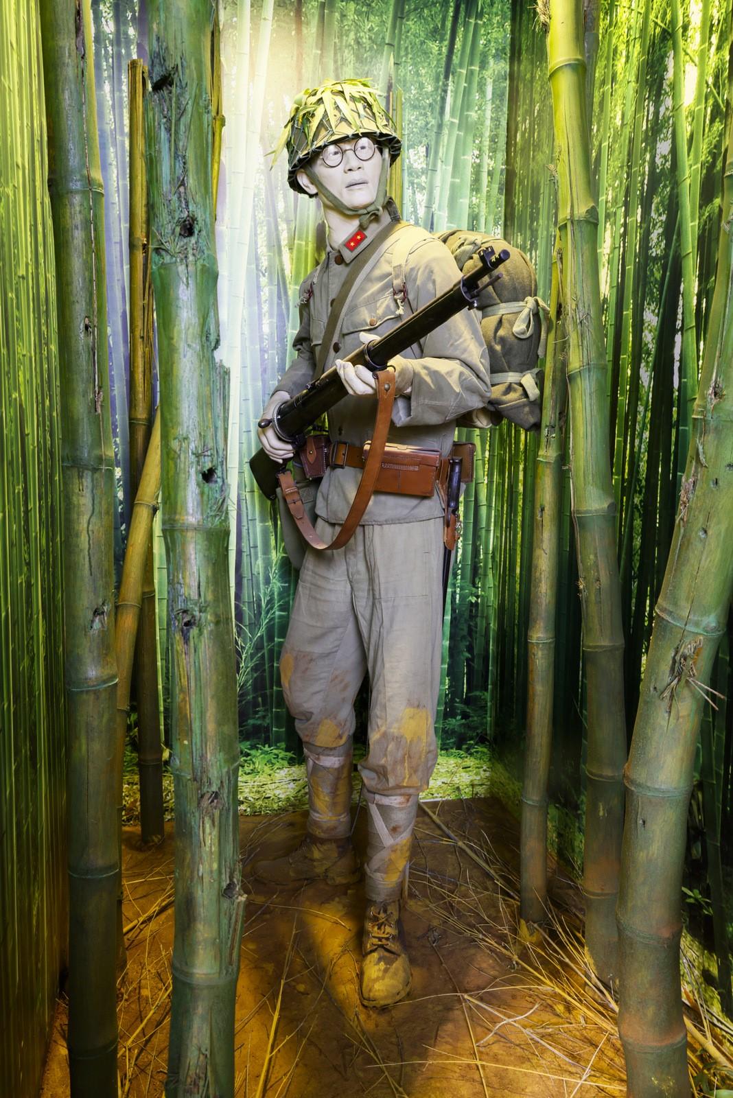 Mannequin of a WW2 Japanese soldier dressed in combat gear, standing in a dense bamboo forest setting. The figure is wearing a helmet, holding a rifle, and carrying a backpack, evoking a wartime jungle environment.