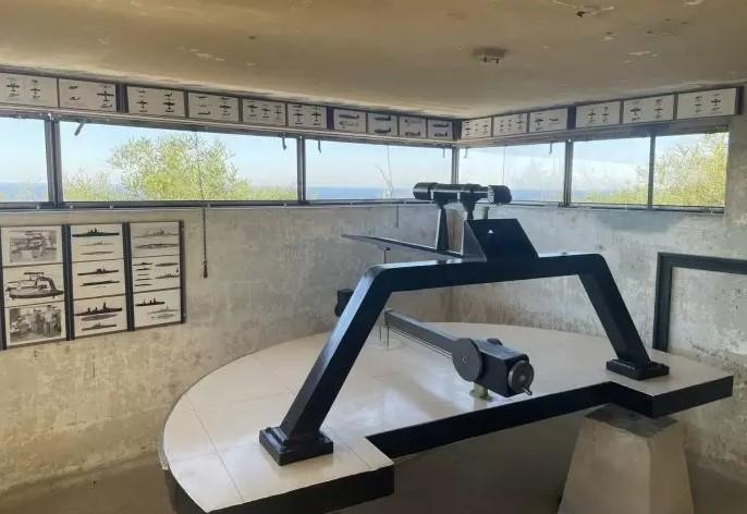 A historical display inside a concrete observation bunker featuring a mounted optical rangefinder or targeting device on a round table (replica Depression Position Finder). The walls have informational posters and diagrams, and windows provide views of the surrounding landscape