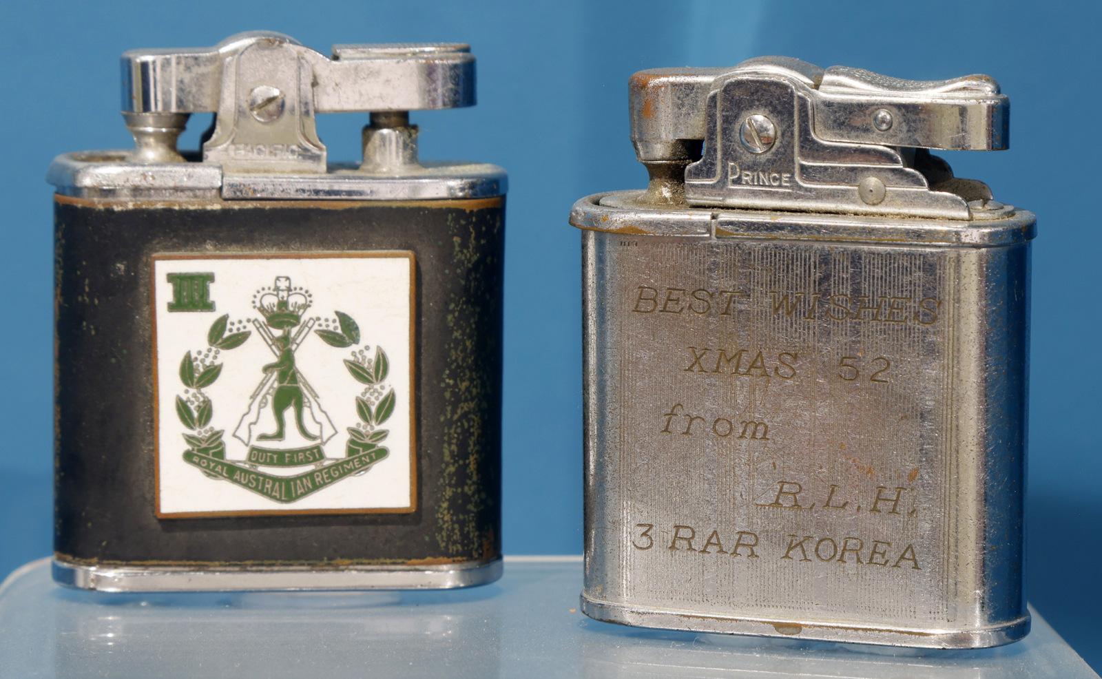 Two vintage metal lighters are displayed side by side. The lighter on the left has a black body with a white panel showing a green emblem of a figure holding scales and a sword, surrounded by laurel branches and a Roman numeral "II" above. The lighter on the right has an engraved silver body with faint text, which appears to commemorate a specific event or person, though the inscription is worn and partially illegible. Both lighters have a flip-top ignition mechanism and reflect a polished, aged appearance.