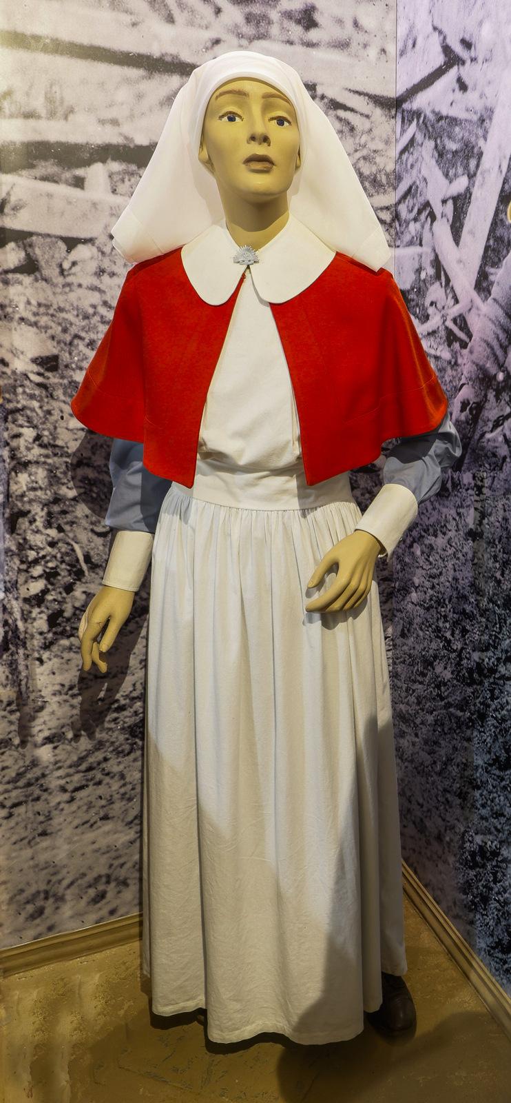 The image shows a mannequin dressed in a World War 1, Australian Army Nursing Service nurse's uniform, likely from the early 20th century. The attire consists of a long white dress, a white apron, and a bright red cape with a rounded white collar. The mannequin also wears a white head covering that drapes over the shoulders. The background features a black-and-white image, depicting a wartime or medical scene.