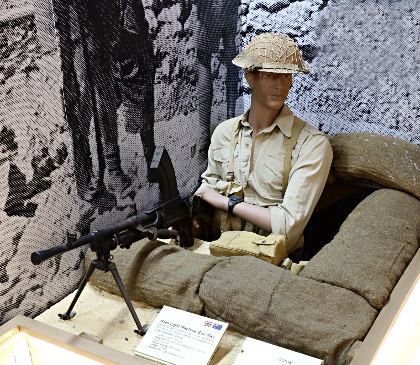 A museum exhibit showing a mannequin dressed in a World War 2 khaki Australian Army military uniform, seated in a sandbag trench. The mannequin is wearing a helmet and is positioned next to a mounted Bren gun. The background features a black-and-white image of soldiers, enhancing the historical wartime setting. Informational plaques are displayed near the exhibit.