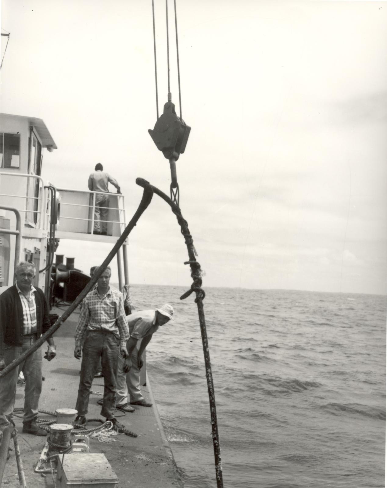 Black and white photograph of a group of men standing on the deck of a ship at sea, retreiving damaged submarine telephone cable to Rottnest Island cable being hoisted by a crane. The horizon and open ocean stretch in the background under a cloudy sky.