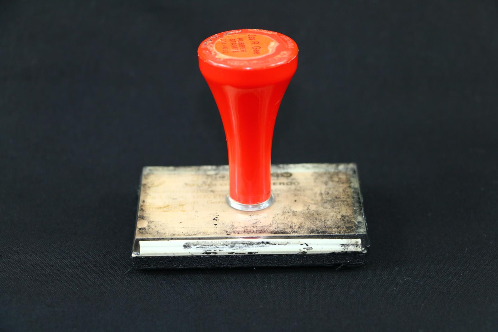 A clear Perspex stamp with red handle. A view from the side The stamp has black ink on the top. The rubber stamp at the bottom is a bit worn.  