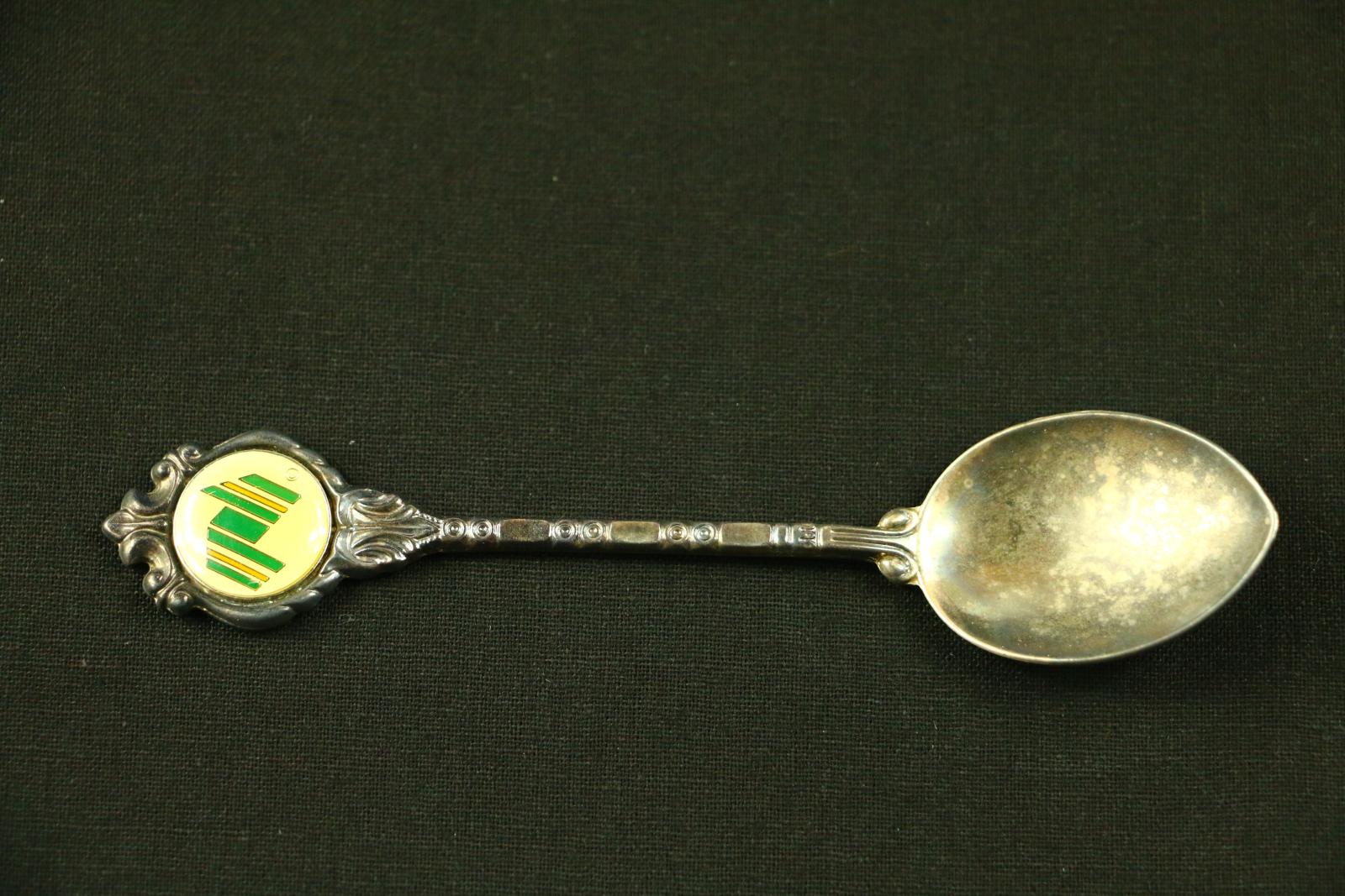 A small, long, thin, silver, brass and enamel 'Stuart' Australian Bicentenary teaspoon. The bowl of the spoon tapers around a point at the front with a shell pattern on the reverse side at the beginning of the stem to the bowl. The round handle of the spoon is intricately decorated around a white, green, gold and yellow enamel badge. On the lower right-hand corner is a very small gold circle outlined with a 'C' in the centre which is also outlined in gold. 