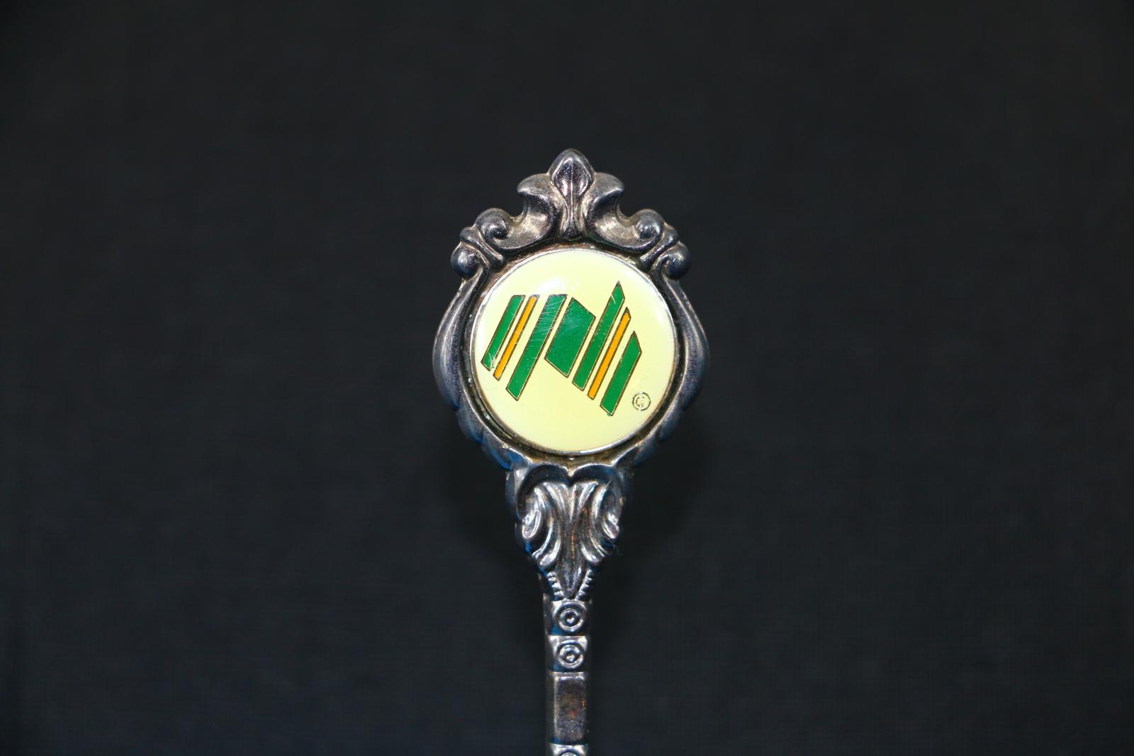 A small, long, thin, silver, brass and enamel 'Stuart' Australian Bicentenary teaspoon. The bowl of the spoon tapers around a point at the front with a shell pattern on the reverse side at the beginning of the stem to the bowl. The round handle of the spoon is intricately decorated around a white, green, gold and yellow enamel badge. On the lower right-hand corner is a very small gold circle outlined with a 'C' in the centre which is also outlined in gold. 