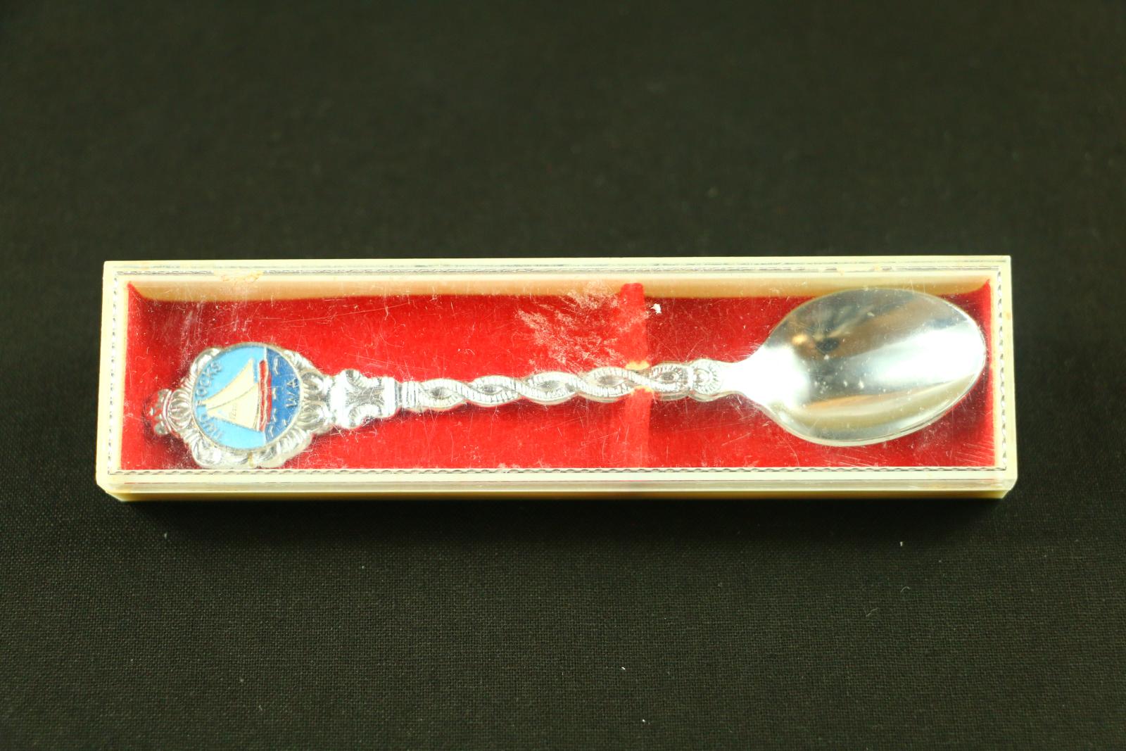 Silver metal, Two Rocks teaspoon. The handle has a twisted pattern with an enamelled logo at top of handle with words 'Two Rocks WA' around picture of 12m sail boat on sea of water. The reverse of the spoon is flat with no embellishments. The spoon comes in a long, white, plastic box. The box has some staining on the underside. A red brushed piece of cardboard to support spoon sits in base of plastic container and there is a long, clear plastic lid which fits the container.