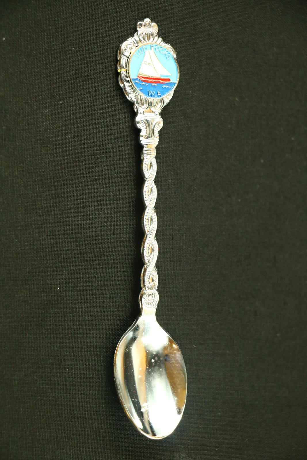 Silver metal, Two Rocks teaspoon. The handle has a twisted pattern with an enamelled logo at top of handle with words 'Two Rocks WA' around picture of 12m sail boat on sea of water. The reverse of the spoon is flat with no embellishments. The spoon comes in a long, white, plastic box. The box has some staining on the underside. A red brushed piece of cardboard to support spoon sits in base of plastic container and there is a long, clear plastic lid which fits the container.