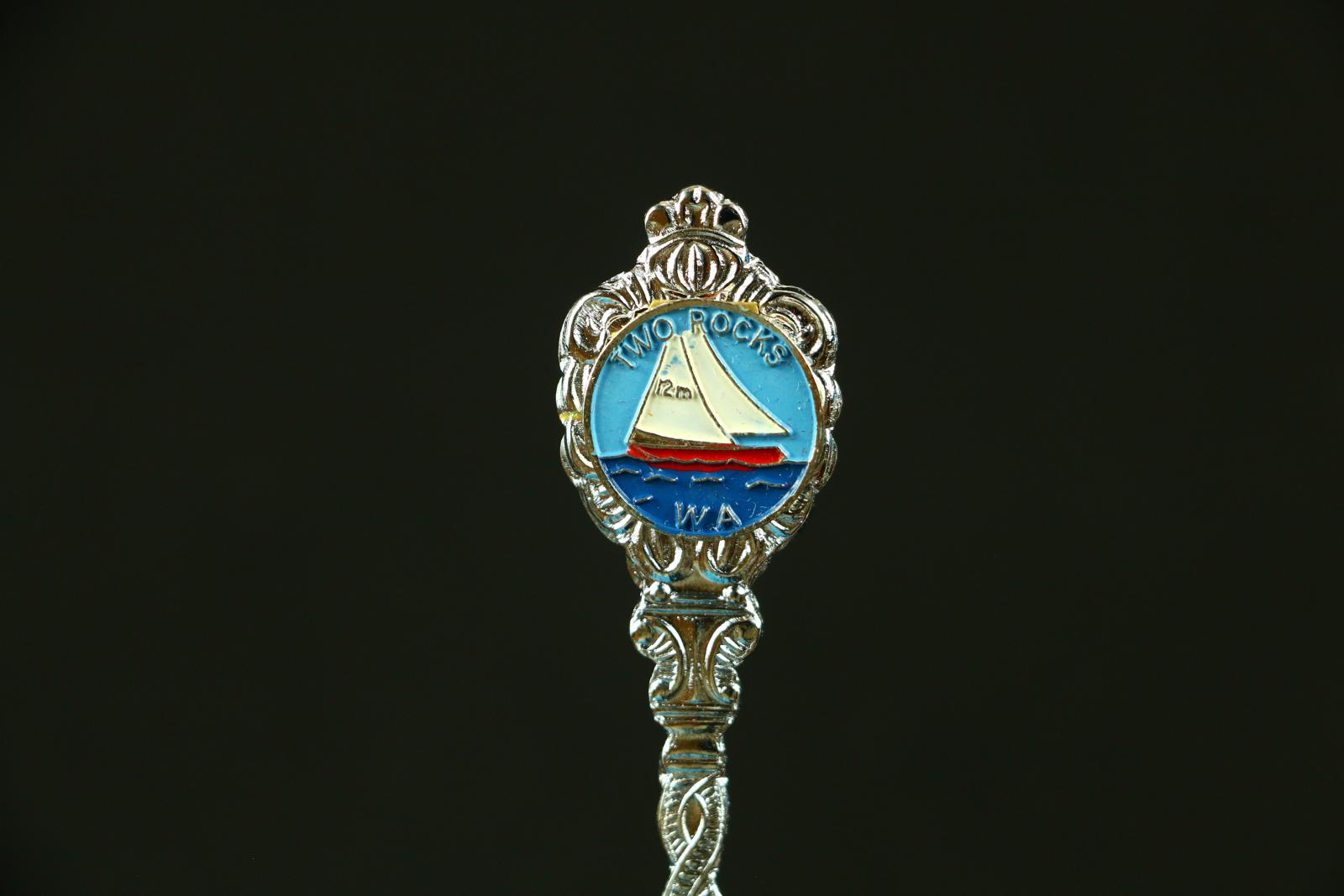 Silver metal, Two Rocks teaspoon. The handle has a twisted pattern with an enamelled logo at top of handle with words 'Two Rocks WA' around picture of 12m sail boat on sea of water. The reverse of the spoon is flat with no embellishments. The spoon comes in a long, white, plastic box. The box has some staining on the underside. A red brushed piece of cardboard to support spoon sits in base of plastic container and there is a long, clear plastic lid which fits the container.