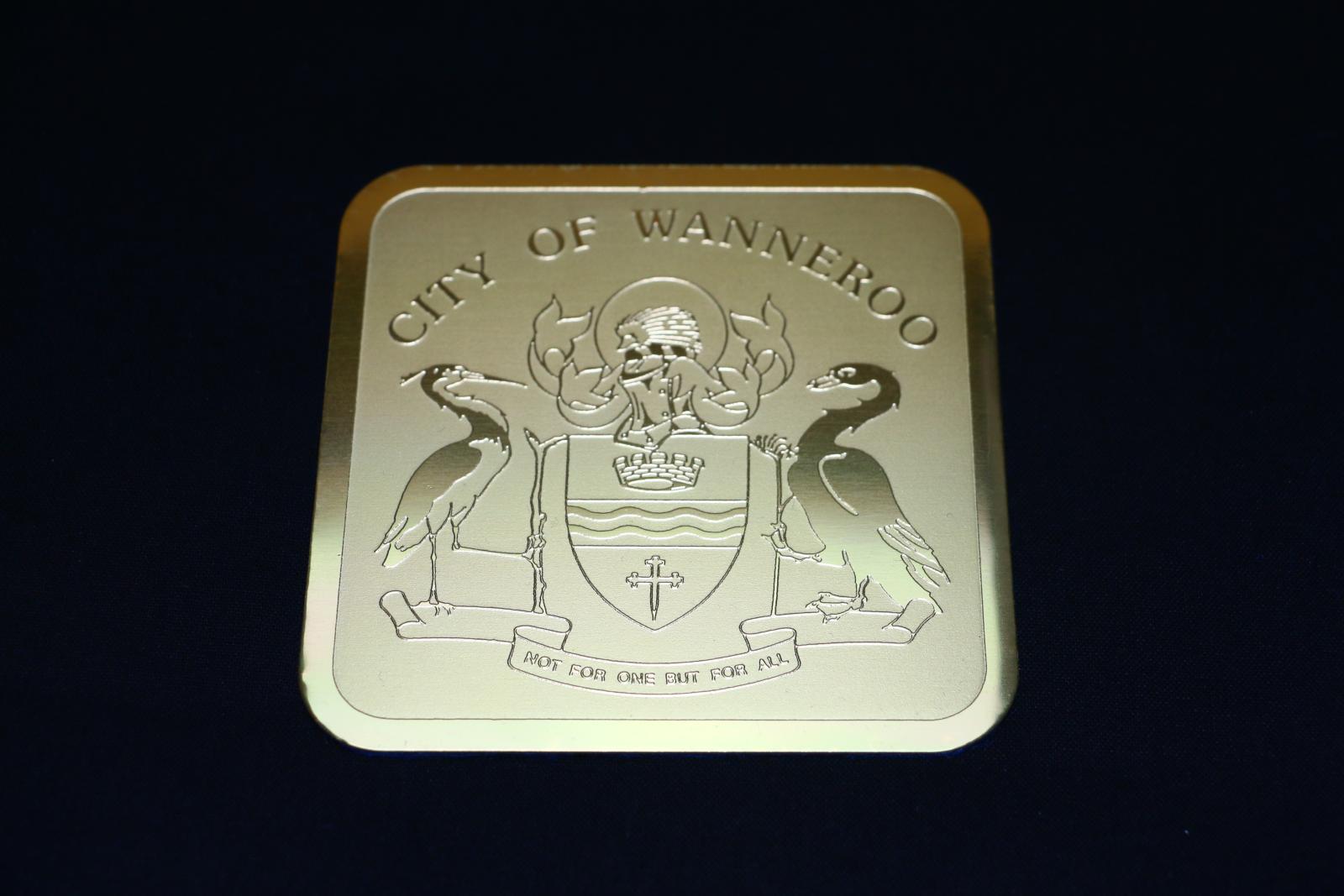 Six identical, square, gold metal, City of Wanneroo coasters. Thin gold metal square with rounded edges. On the front is a thin shiny gold border with the matte gold centre. Written across the top in raised, shiny gold letters is 'CITY OF WANNEROO'. Underneath, covering the majority of the coaster, is a raised, shiny gold, stylised crest with two tall water birds holding a shield between them with the helmet and ribbon motif on top. Underneath the shield is a small scroll with 'NOT FOR ONE BUT FOR ALL'