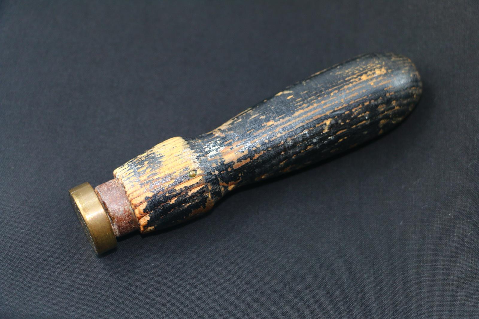 A brown wooden handle, painted black, wax seal that is shaped for hand use. The paint on the wooden handle is peeling away. A brass screw is located on one side of the wooden handle. A solid brass circular seal with stamp imprint is located at one end of the object.