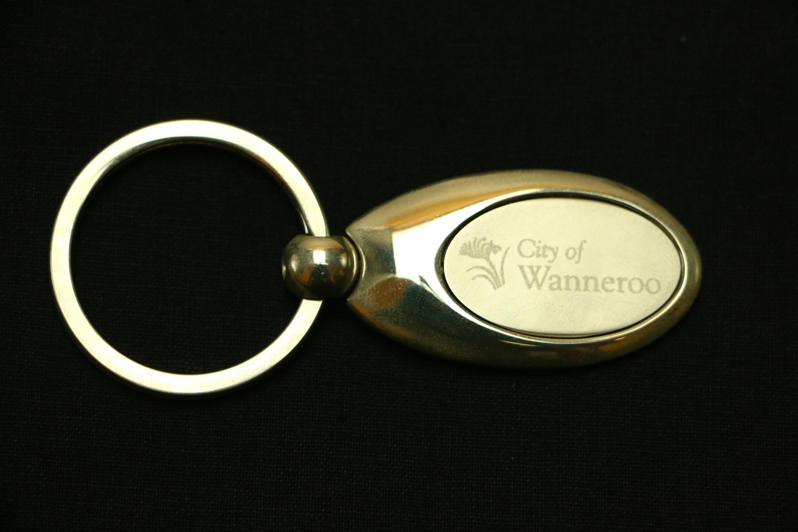 Oval, shiny, silver coloured, rounded metal disc, branded with City of Wanneroo insignia. One end of disc is connected to rotating spherical link, through which a large, flat, split-ring is threaded. Disc has a flat, matte, oval-shaped insert with official branding printed in silver onto the metal, which reads 'City of / Wanneroo' (on right) with a kangaroo paw (on left).