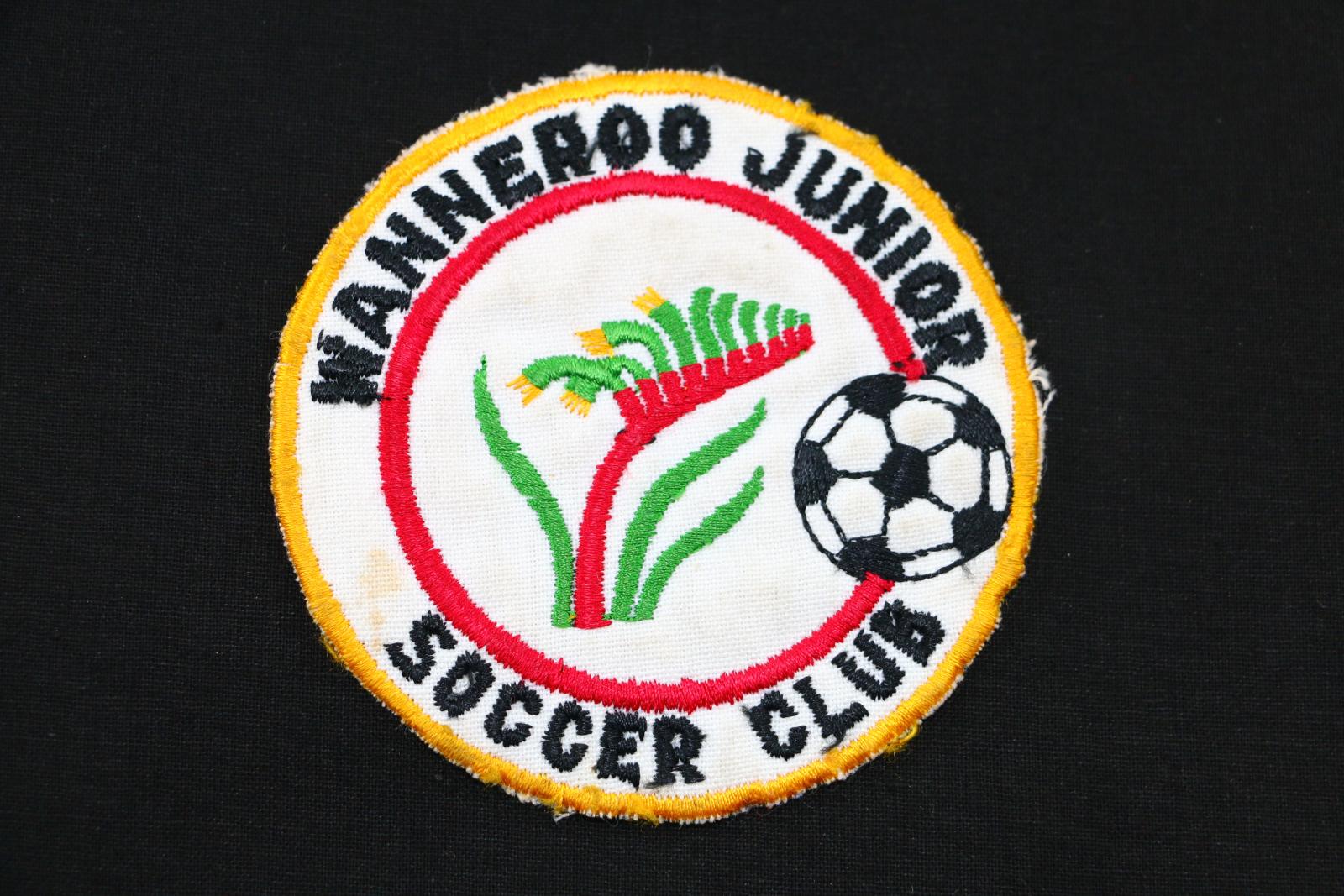 Thin, flat, circular white cloth patch with coloured machine embroidered insignia reading 'WANNEROO JUNIOR / SOCCER CLUB'. Patch has golden outer edges with black text around top and bottom of border, black and white soccer ball on right hand side, and central red circle enclosing a red, green, and yellow stylised kangaroo paw. Back of patch is covered in semi-sheer cream coloured felt-like fabric. 