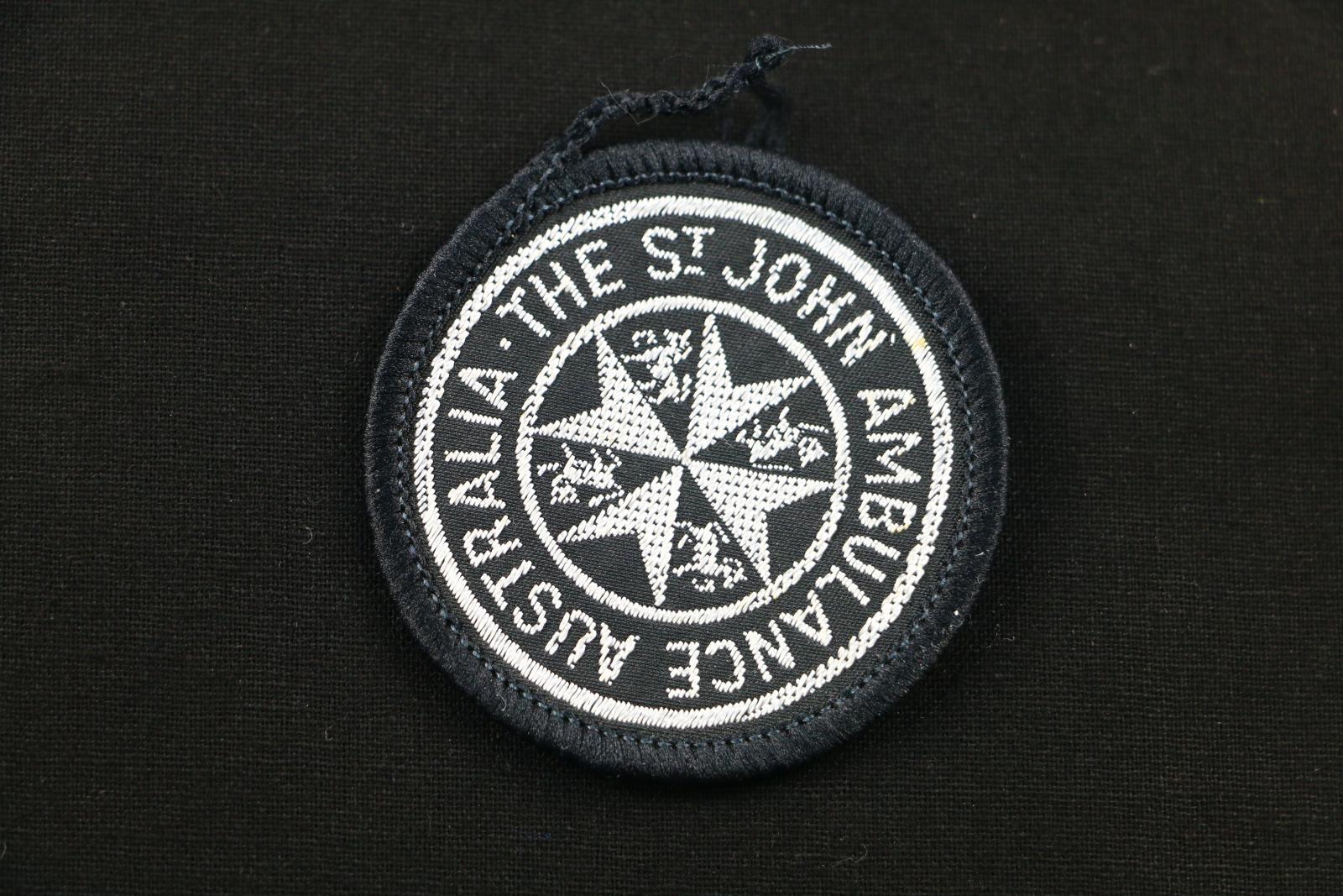 Circular, black cloth badge with white stitched lettering and logo. The badge is black and has the St John Ambulance logo with the following text around the border 'THE ST JOHN AMBULANCE BRIGADE' There is black stitching around the outside edge of the badge.