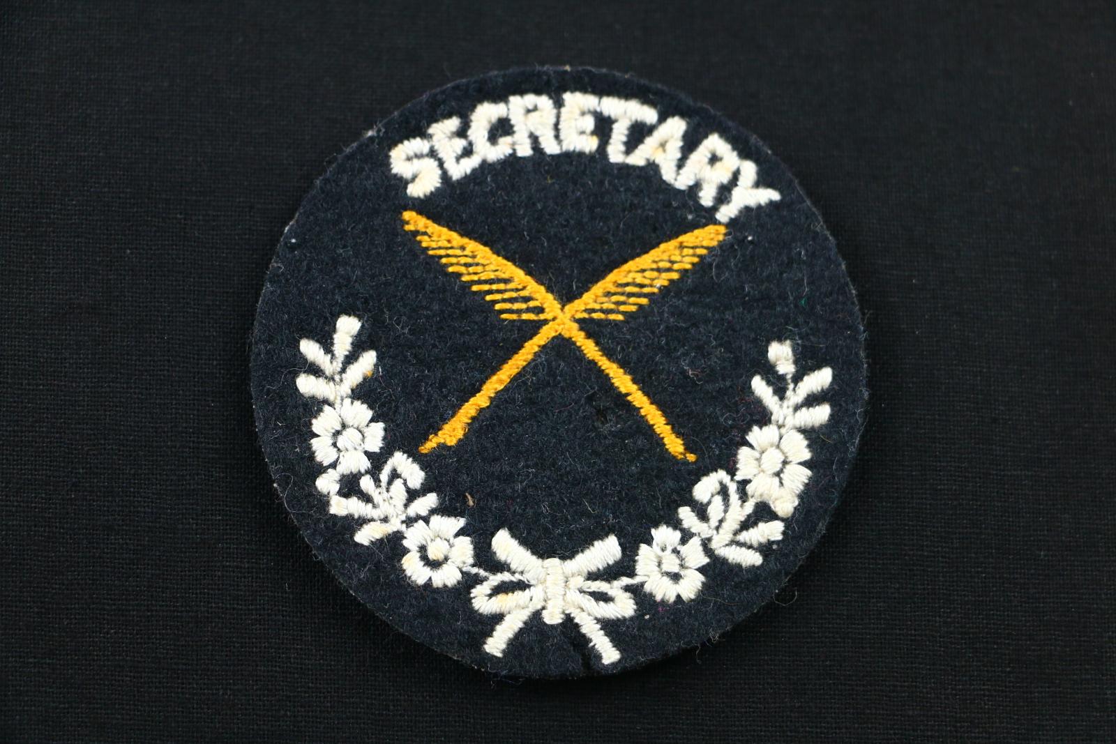 Circular, black cloth badge with yellow and white stitching. The badge has white text 'SECRETARY' at the top. Underneath the text is a yellow stitched design of two diagonally overlapping feather quill pens. Under that is a white stitched design of flowers and and a bow in an arch shape.