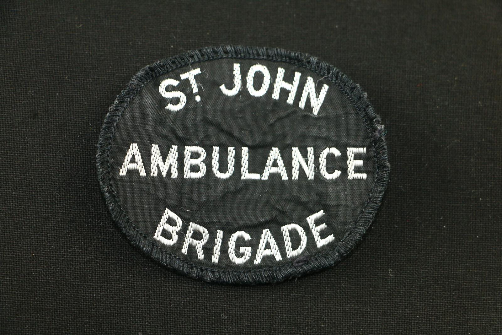 Oval shaped black cloth badge with white stitched lettering. The badge is black with large text 'ST JOHN/ AMBULANCE/ BRIGADE' The badge has a stitched black border.