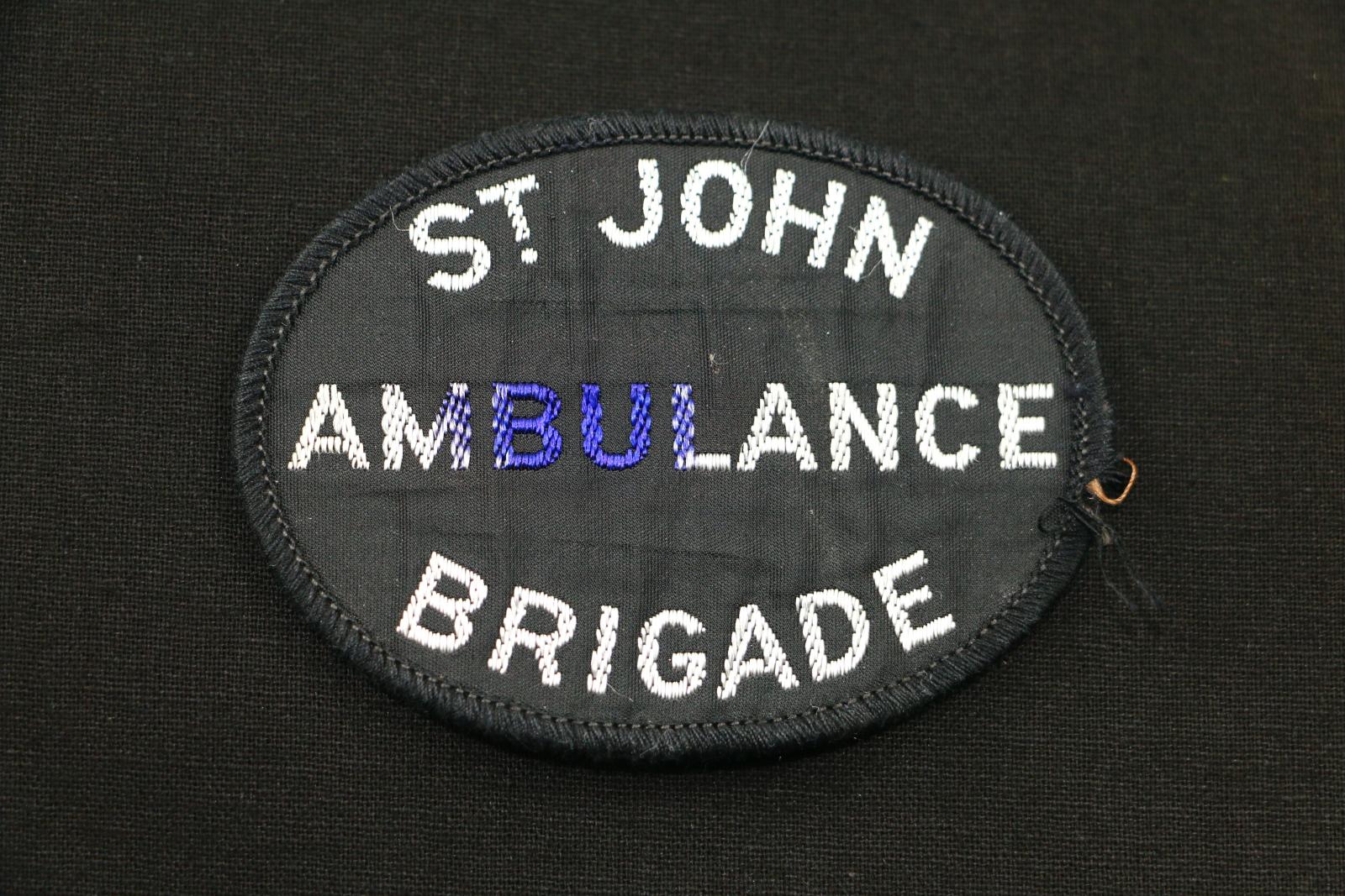 Oval shaped black cloth badge with white stitched lettering.  The letters 'M,B,U, and L have blue ink marks on them. The badge is black with large text 'ST JOHN/ AMBULANCE/ BRIGADE' The badge has a stitched black border.