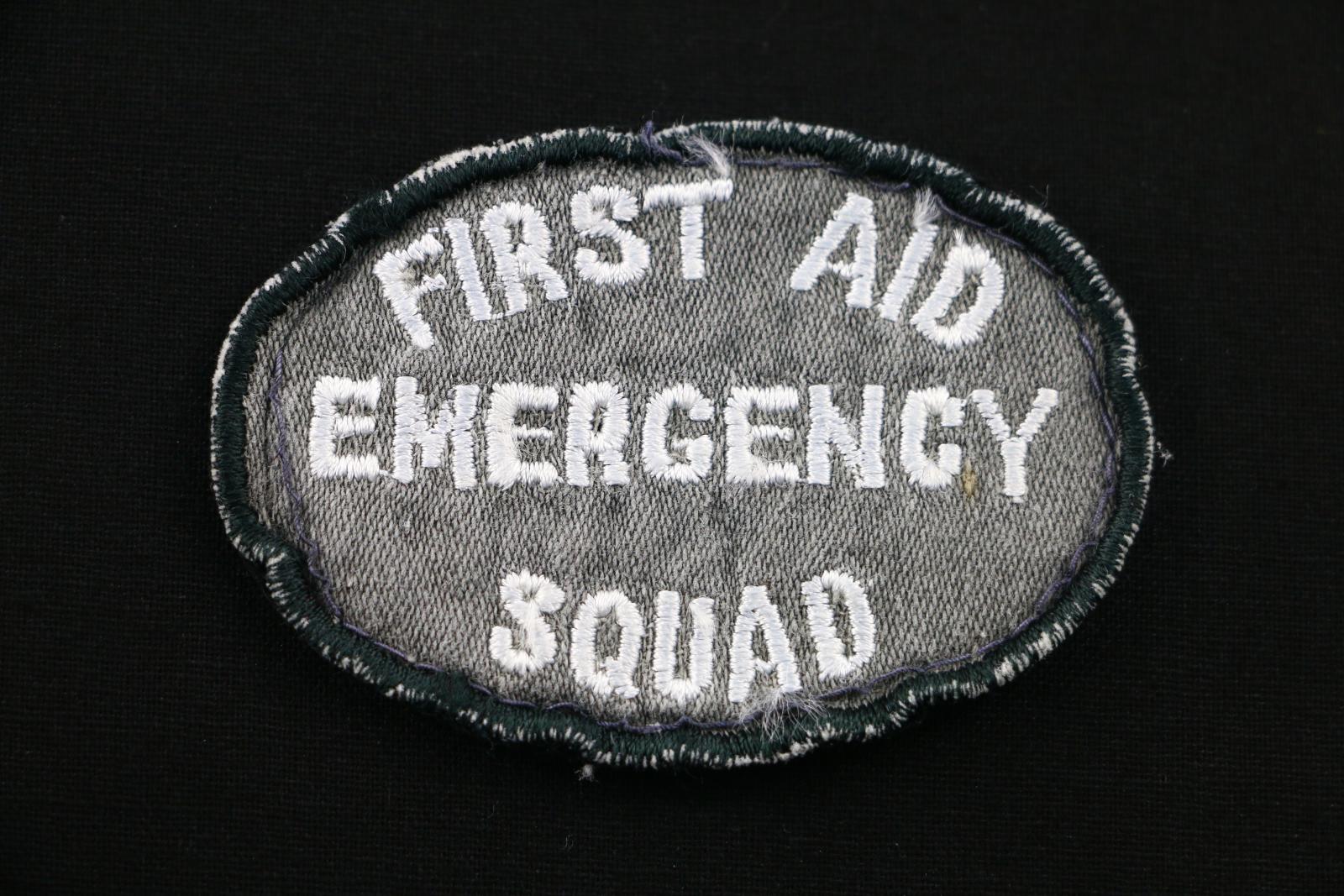 Oval, shaped grey, black and white cloth badge. The badge is grey with a black stitched border and white stitched lettering. The text is 'FIRST AID/ EMERGENCY SQUAD' . The back of the badge is black.