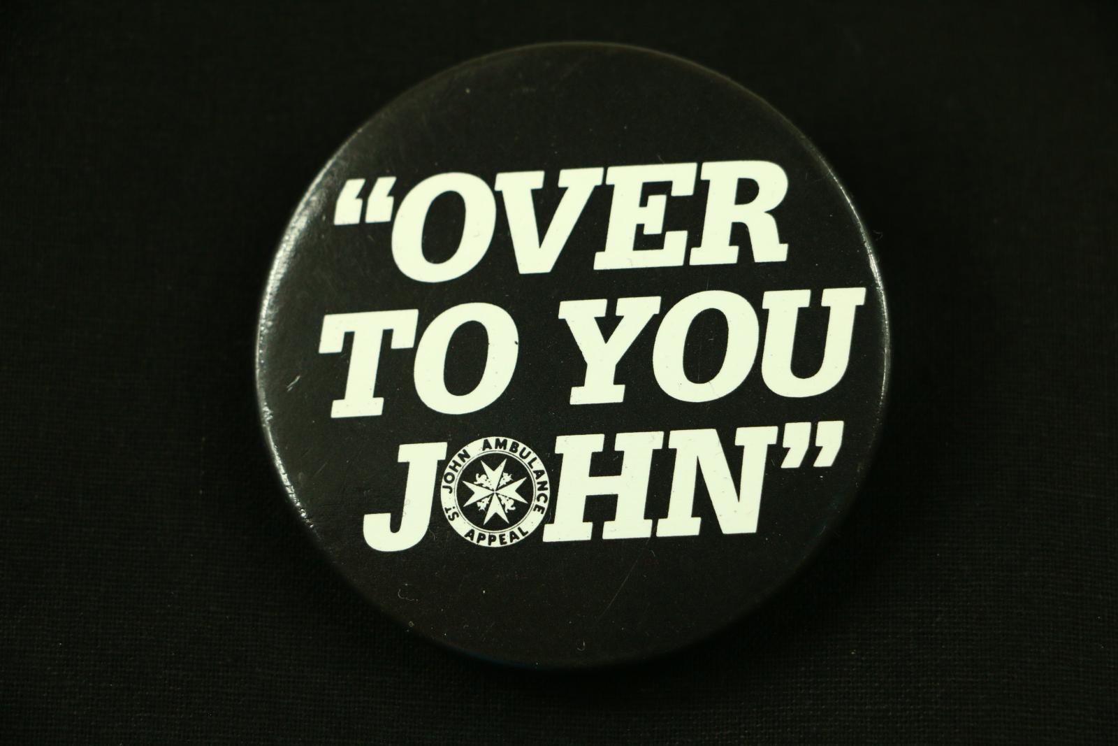 Circular black and white metal badge with plastic coating. The badge has a black background with white lettering. The text is as follows ' ''OVER/TO YOU/ JOHN'' ' The letter O in the word John has been replaced with the St John Ambulance logo. The text around the border of the logo is 'ST JOHN AMBULANCE APPEAL'